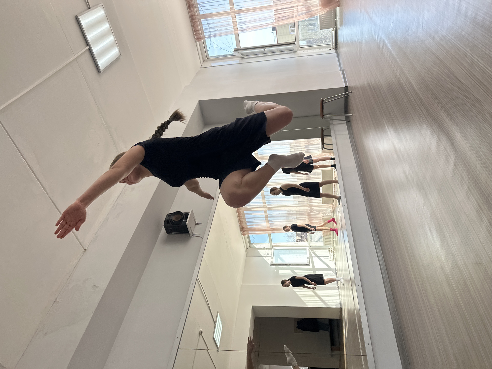 The photo captures a dance studio in Votkinsk, Russia. A young woman with a braided ponytail is suspended mid-air, performing a dance move. She is wearing a black top, black shorts, and white socks. Her right arm is extended to the left, and her legs are positioned to the right. The background features a mirror reflecting several dancers in similar attire, practicing along the wall. There is a window with curtains, a set of chairs along the mirror, a light fixture on the ceiling, and a large spotlight on the wall. The wall is a plain white with visible marks. The studio floor is a light brown textured surface. The room has a high ceiling, giving it a spacious appearance.