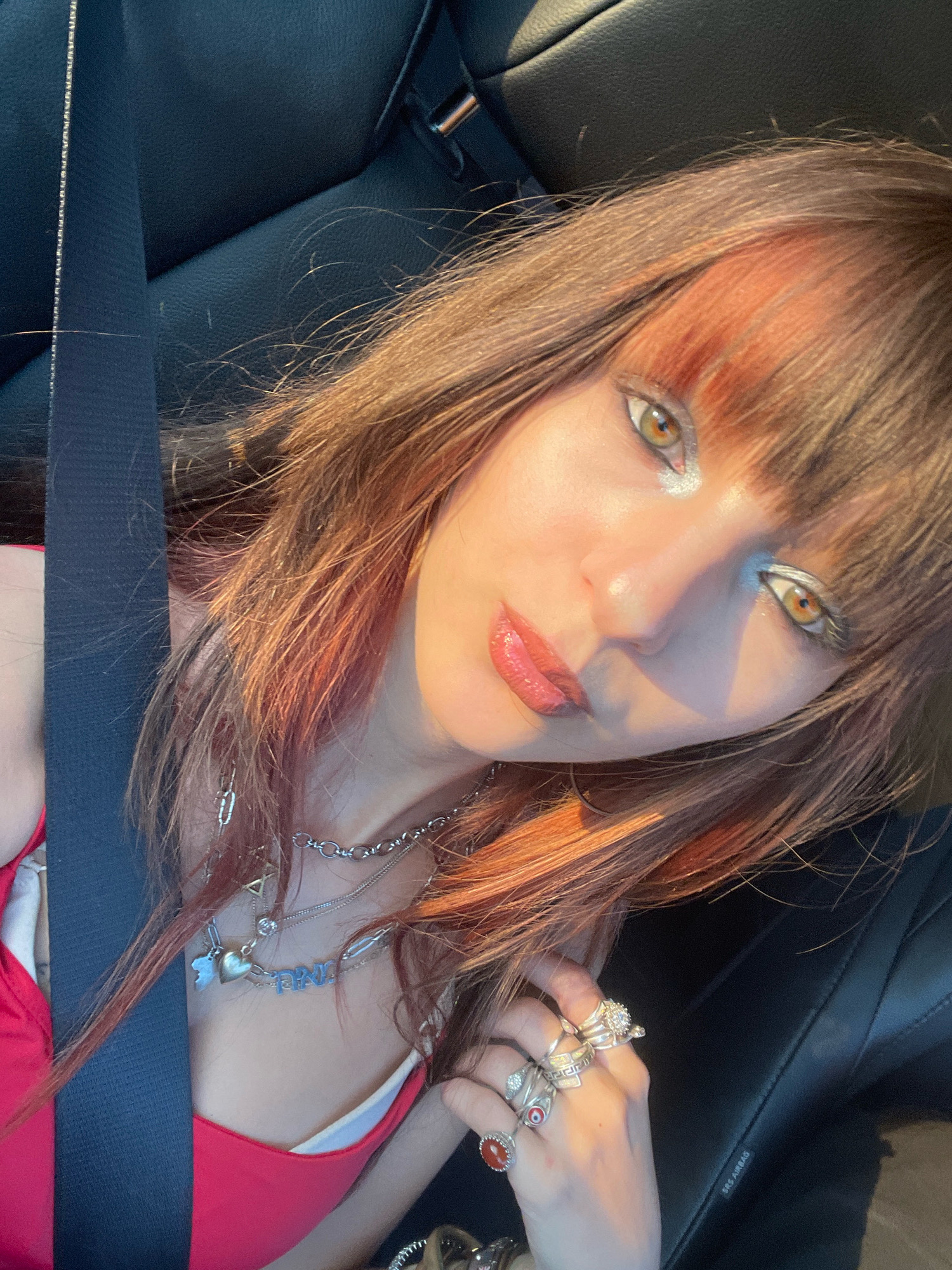A young woman with auburn hair and bangs is taking a selfie in a car. She is wearing a red top and multiple necklaces, including one with a heart pendant and another spelling out "PINK". She has rings on her fingers. The background shows the black interior of a car, including a seatbelt. The lighting suggests it is daytime, possibly late afternoon, with sunlight casting shadows. The location is not determinable from the image.