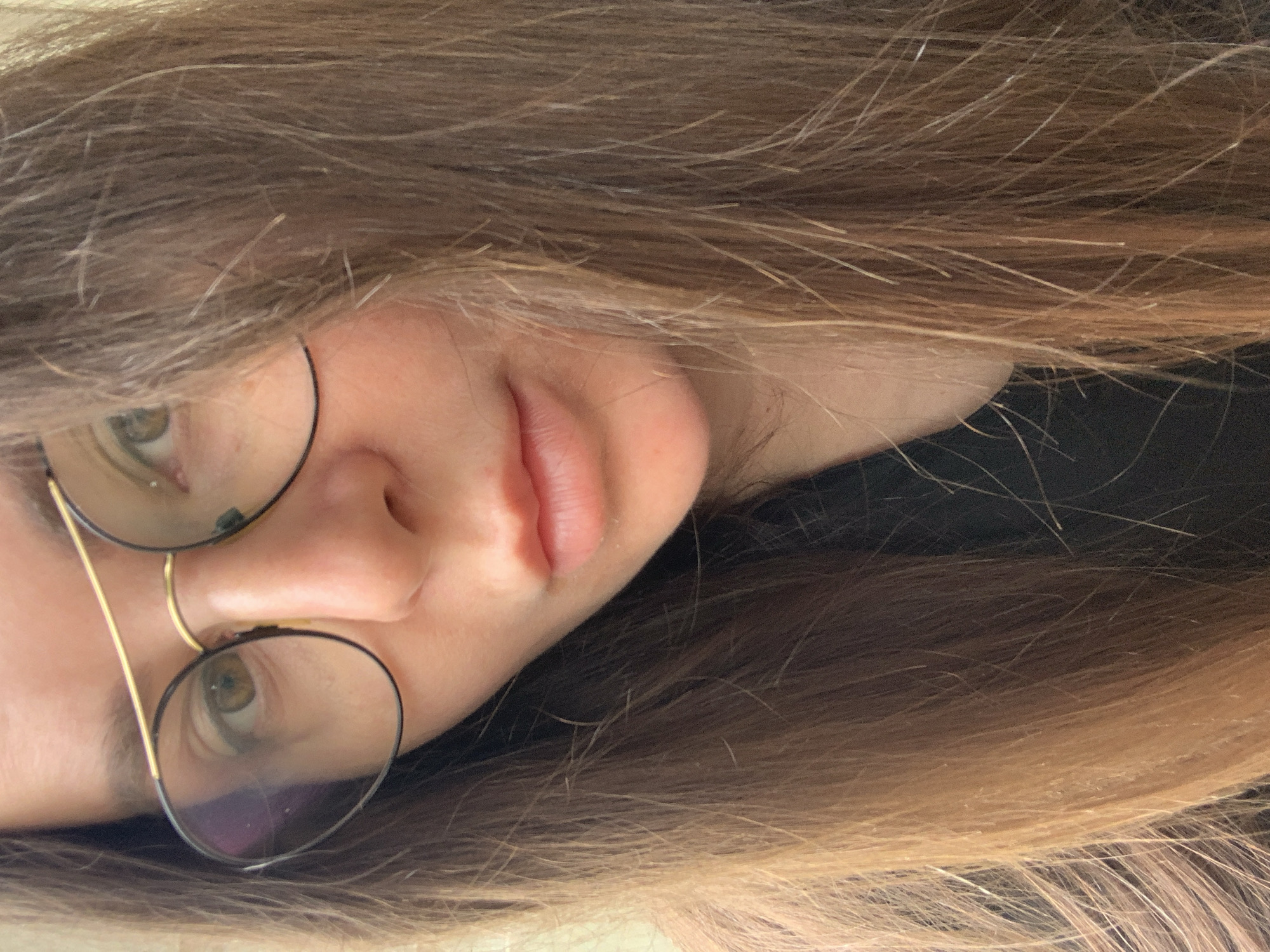 A close-up, slightly distorted, portrait shot of a person's face. The person has fair skin, light brown hair with some lighter highlights, and is wearing round, wire-rimmed glasses. The camera angle is slightly angled, giving a unique perspective of the facial features. The background is filled with the person's hair, with some areas appearing darker. The person is not in any city.