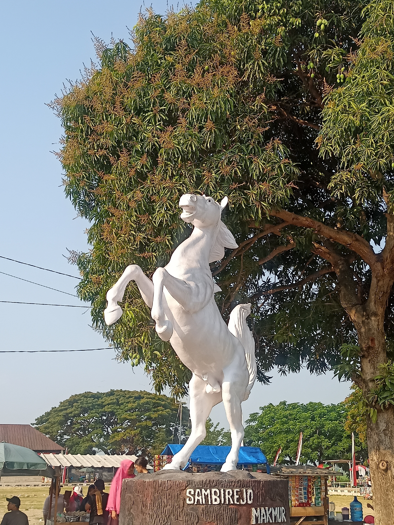A white horse statue stands on a wooden base inscribed with "SAMBIREJO MAKMUR" in the city of Ngrompongan, Indonesia. The statue is positioned in front of a large, leafy tree with visible mangoes and flowers. In the background, there are several market stalls with people browsing and vendors attending to their businesses. A few red and white flags are also visible, suggesting a local event or cultural display. The sky is clear and blue.