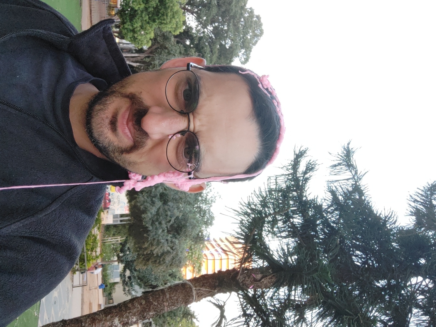 This image features a close-up portrait of an adult man with dark hair, a beard, and round, dark-framed glasses. He wears a dark, possibly black, fleece jacket. His mouth is open, suggesting an expression of surprise or amusement, with his eyes looking directly towards the viewer. Pink, stringy foam or similar playful material is splattered across his forehead, hair, and beard, with a long strand hanging down from his face.
The background depicts an outdoor setting, likely a park or urban green space. Various trees, including a prominent conifer with dark green needles in the lower right, and other leafy trees, are visible. In the middle ground, faint details of urban structures, possibly buildings and a playground with colorful elements, can be seen, along with what appears to be a paved area and a grassy patch. The lighting indicates daytime, possibly a bright but not harshly sunny day. The overall atmosphere appears lighthearted and spontaneous, capturing a playful moment. No visible text is present. The scene is set in Tel-Aviv, Israel.
