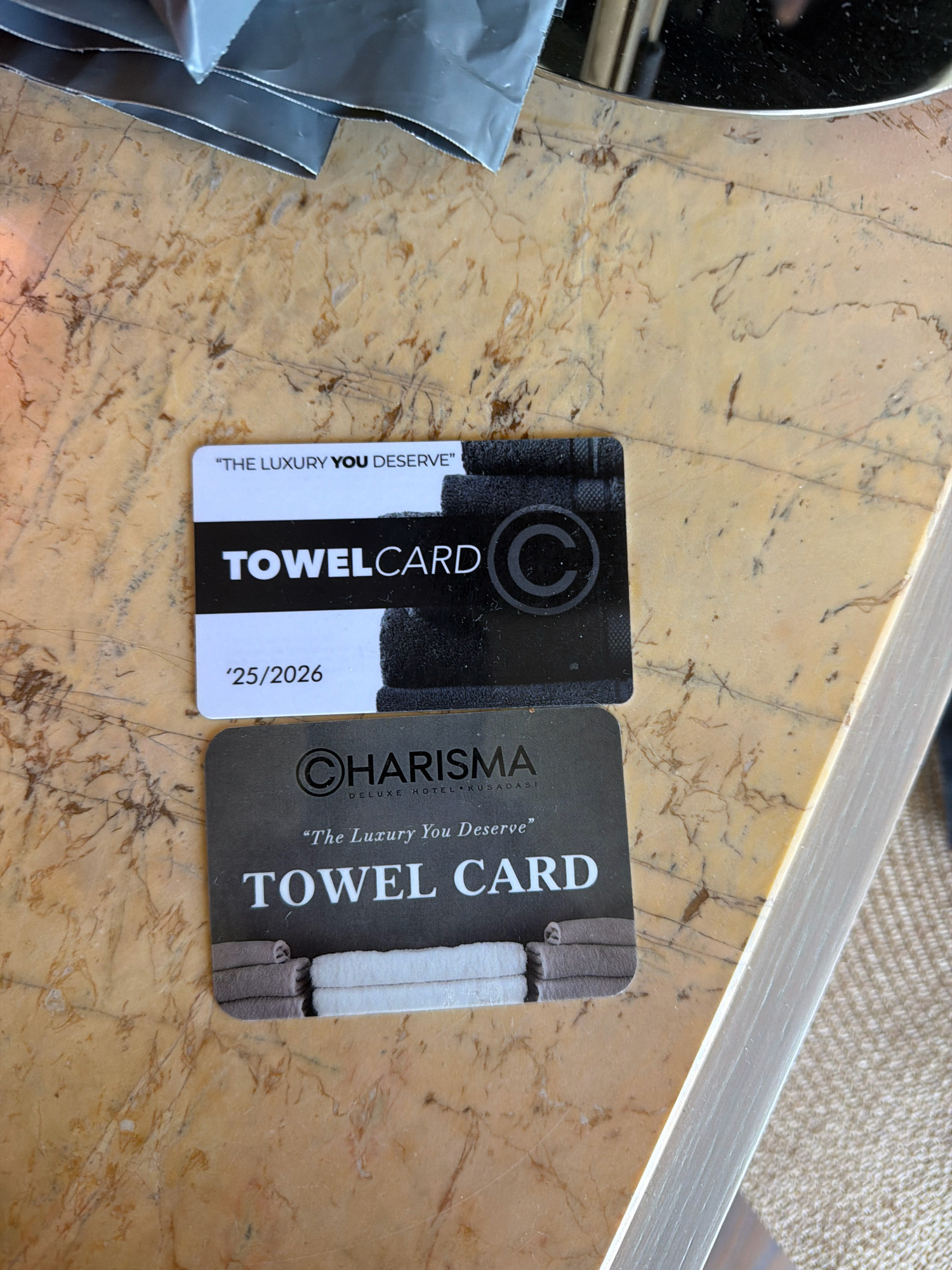 The image shows two "Towel Card" items resting on a marble-like surface. The top card is white with black text and a black band featuring the word "TOWELCARD" and a circular logo with a "C". Below this text, there is a date "'25/2026". The bottom card is dark gray with white text. It features a circular logo with a "C" and the word "CHARISMA" in larger font, with smaller text below that reads "DELUXE HOTEL KUŞADASI". Below "CHARISMA", the phrase "The Luxury You Deserve" is written, followed by "TOWEL CARD" in large letters. This bottom card also has an image of neatly folded towels at the bottom. The overall scene suggests a hotel amenity or promotional item from Kuşadası, Turkey.