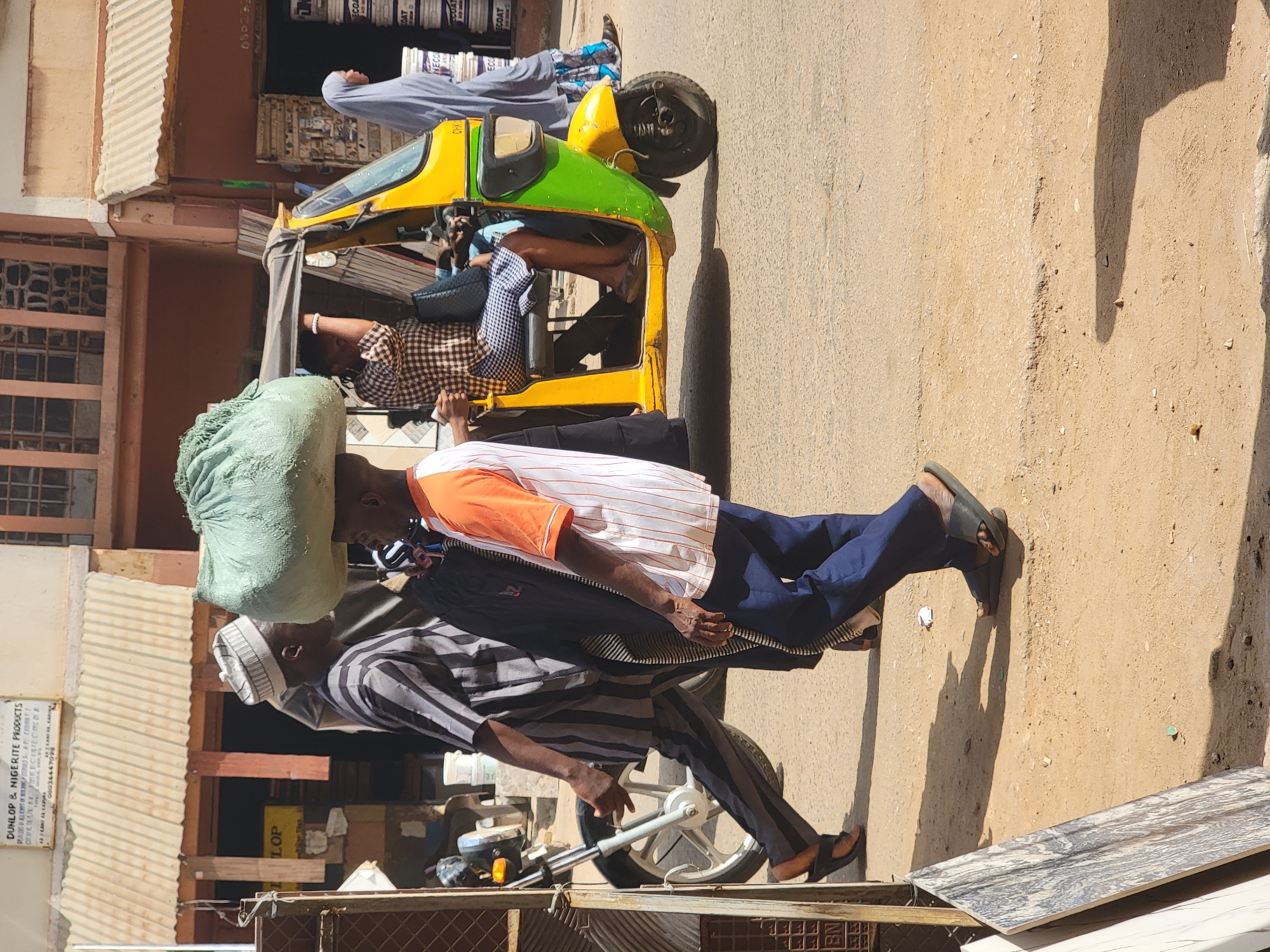 The image shows a street scene in Maiburiji, Nigeria. Two men are carrying a large green sack on their heads, one in front and one behind. The man in front wears a white and grey striped shirt, a white hat, and black pants. The man behind wears a white shirt with orange and brown stripes and blue pants. Both men wear sandals. Behind them is a motorcycle-taxi, known locally as a keke or tricycle. The keke is yellow and green and has passengers inside. The background includes buildings with corrugated metal roofs and a sign that reads "Dunlop & Nigerite Products." The street surface is sandy.