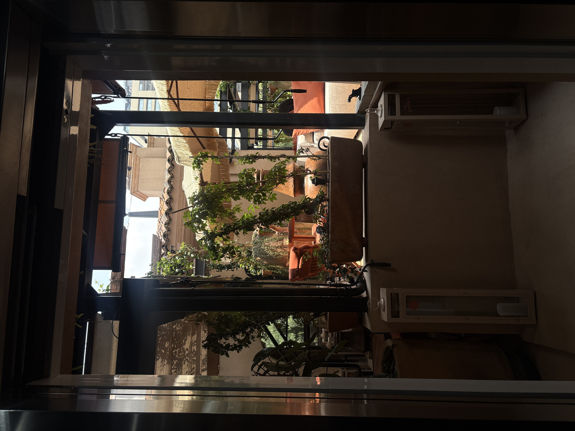 This is a close-up, eye-level shot taken from inside what appears to be an elevator, looking out through its open doors. The view is of an outdoor patio or balcony area with lush greenery and seating. The architecture suggests a Mediterranean or Spanish colonial influence, with terracotta tile roofing visible in the background. Sunlight filters through the leaves, creating dappled patterns on the ground and walls.
On the patio, there's a long, rectangular planter filled with plants, and a round wooden table with an orange chair is positioned behind it. Further back, cushioned seating areas are visible, some with large, umbrella-like structures providing shade. Two tall, slender lanterns, one on either side of the opening, flank the view, each containing a white candle. The lantern on the right has the text "Sky Garden" printed vertically on its side. The overall atmosphere is serene and inviting, hinting at a stylish rooftop bar or restaurant, possibly in a trendy neighborhood of Mexico City. The metallic walls of the elevator frame the scene, creating a sense of transition between indoor and outdoor spaces.