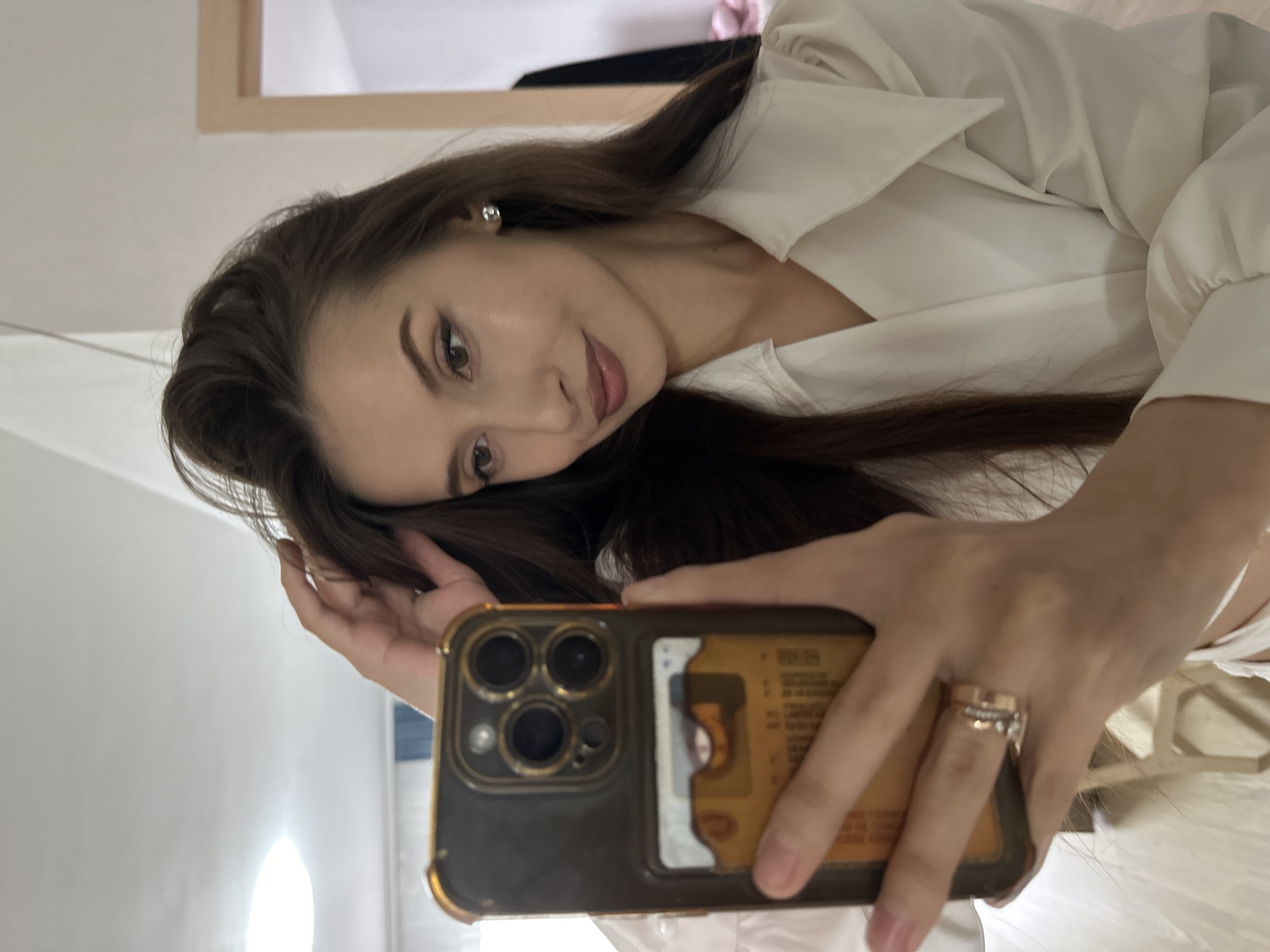 A young woman with long, dark brown hair is depicted taking a mirror selfie indoors. Her head is gently tilted, and she gazes towards the camera with a soft, subtle smile. She wears a white collared shirt with puffed sleeves. A silver stud earring is visible in her left ear, and her eyes are subtly enhanced with makeup.
She holds a smartphone, appearing to be an iPhone with a triple camera system, in her right hand. The phone is protected by a case featuring a gold-toned frame and a clear back panel. Inserted into the clear slot of the case is a yellowish-orange card, which displays partially visible text including "MOCK CREDIT" and a stylized number "5", along with other illegible characters, suggesting it might be a sample or novelty item. On her right hand's ring finger, she wears a stack of rings, including a plain gold band and another adorned with small, sparkling stones.
The background is light-colored, indicating an indoor setting with a pale wall or furniture. A light wooden frame is visible in the upper right quadrant of the image. A bright light source creates a strong reflection in the lower left. The overall atmosphere is serene, with the woman's expression conveying calmness. While the image itself provides no direct geographical cues, the context provided indicates the location is Yekaterinburg, Russia.