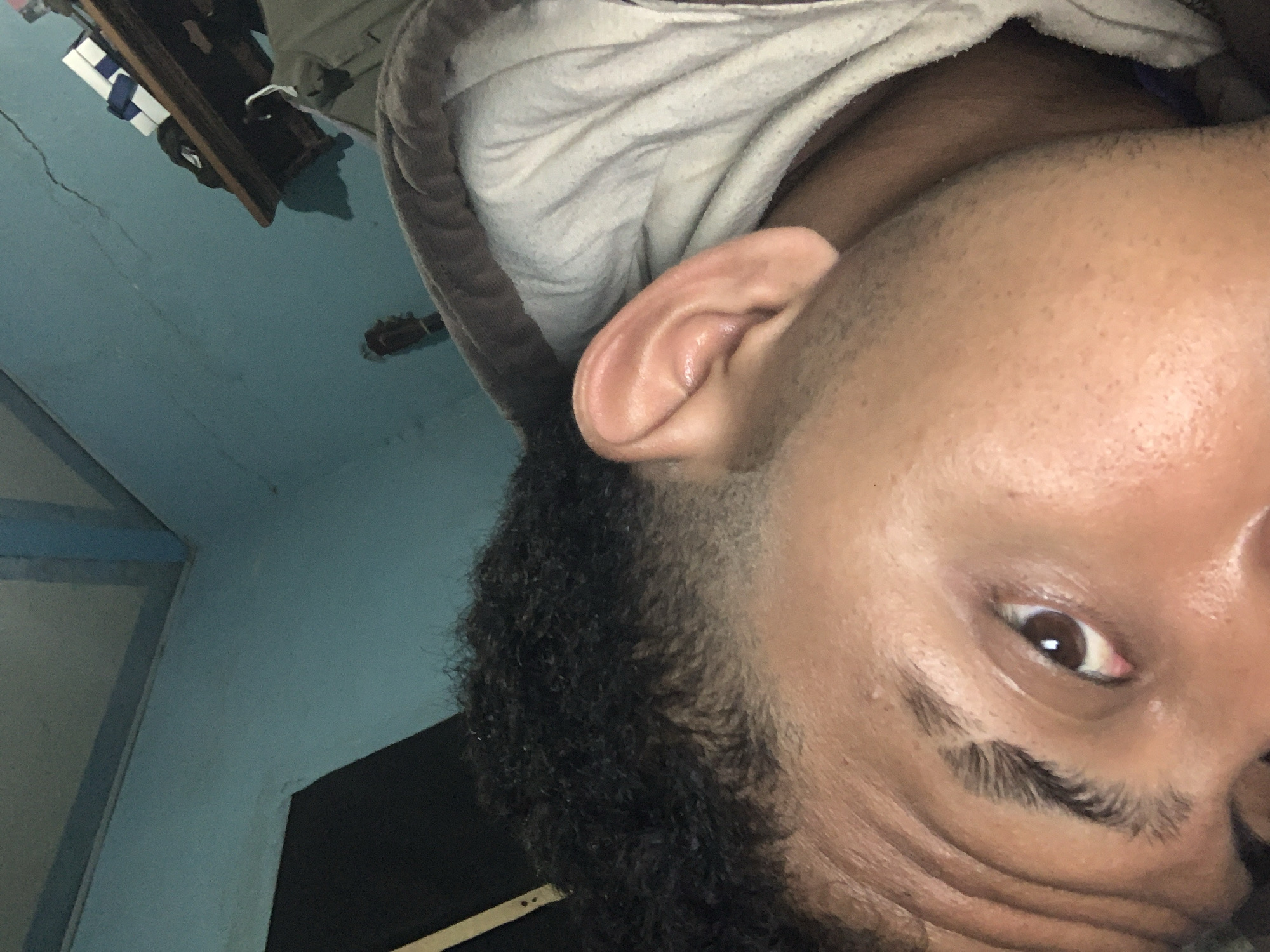 The image, taken from a close-up, low-angle perspective, focuses on a person's face and upper body, oriented such that the head is angled upwards and to the right. The subject appears to be lying down, possibly in a bed, as evidenced by a crumpled white and gray patterned fabric in the background. The person has dark, curly hair and visible facial features including an eye with a brown iris and a pinkish-white sclera, an eyebrow with a distinct shape, and an ear. The skin tone is medium brown. The person is wearing a gray hooded sweatshirt.
The background environment suggests an indoor setting with a blue-painted wall and a ceiling that is also blue. A shelf unit is visible on the right side of the image, containing several objects, including a white box with a blue ribbon. The overall lighting is dim, with shadows present.
The image does not provide any information to determine the specific location within the city of Santa Ana, Venezuela.