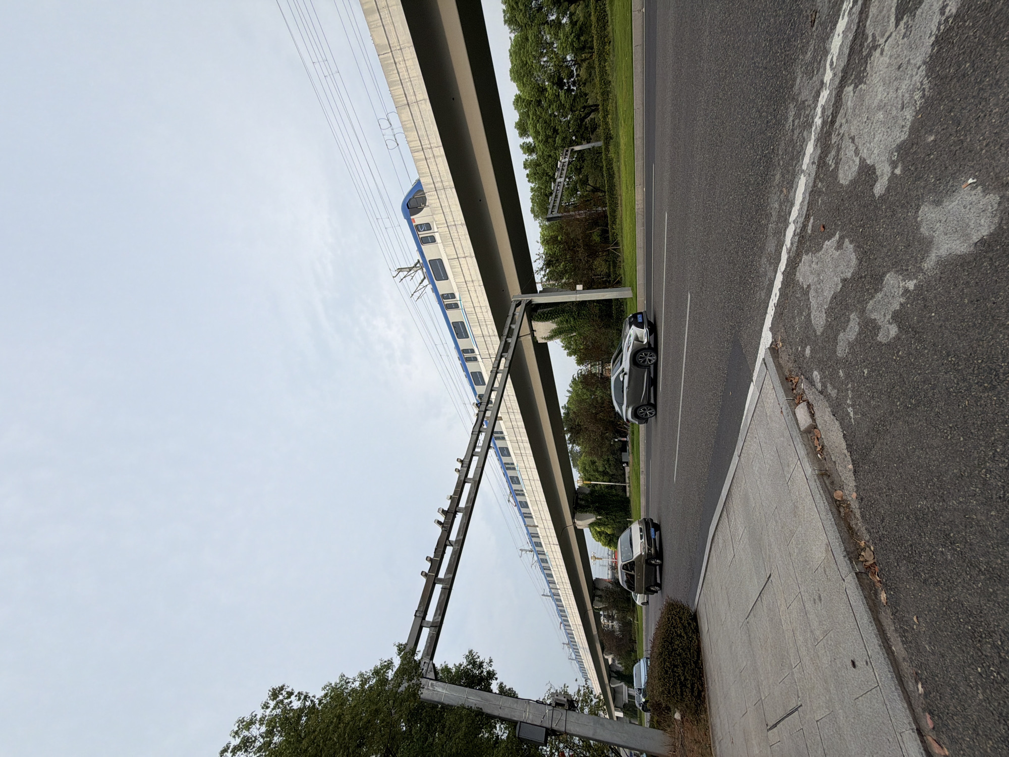 An elevated train line with a blue and white train is visible over a multi-lane road in Jiangjin, China. The train is moving from left to right. Cars are driving on the road below. The road is paved with asphalt and has white lane markings. To the left of the road is a sidewalk made of grey concrete tiles. Trees and green foliage are present behind the road. The sky is overcast.