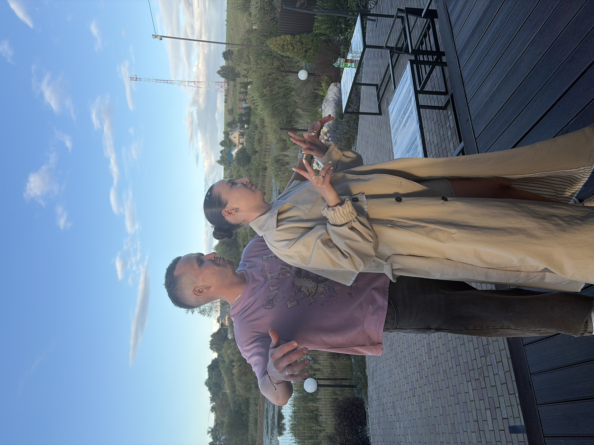 In Karsha, Russia, a man and a woman stand on a wooden deck with a brick patio in the background. The man, wearing a pink t-shirt and dark pants, is gesturing with his left hand, which has a ring on his finger and a bracelet on his wrist. The woman, in a beige trench coat, stands next to him, also gesturing with her hands. Behind them, a body of water is visible, with trees and houses in the distance, under a partly cloudy blue sky. A cell tower stands on a hill in the background. Two tables with chairs are set up on the patio, with drinks on one of them.