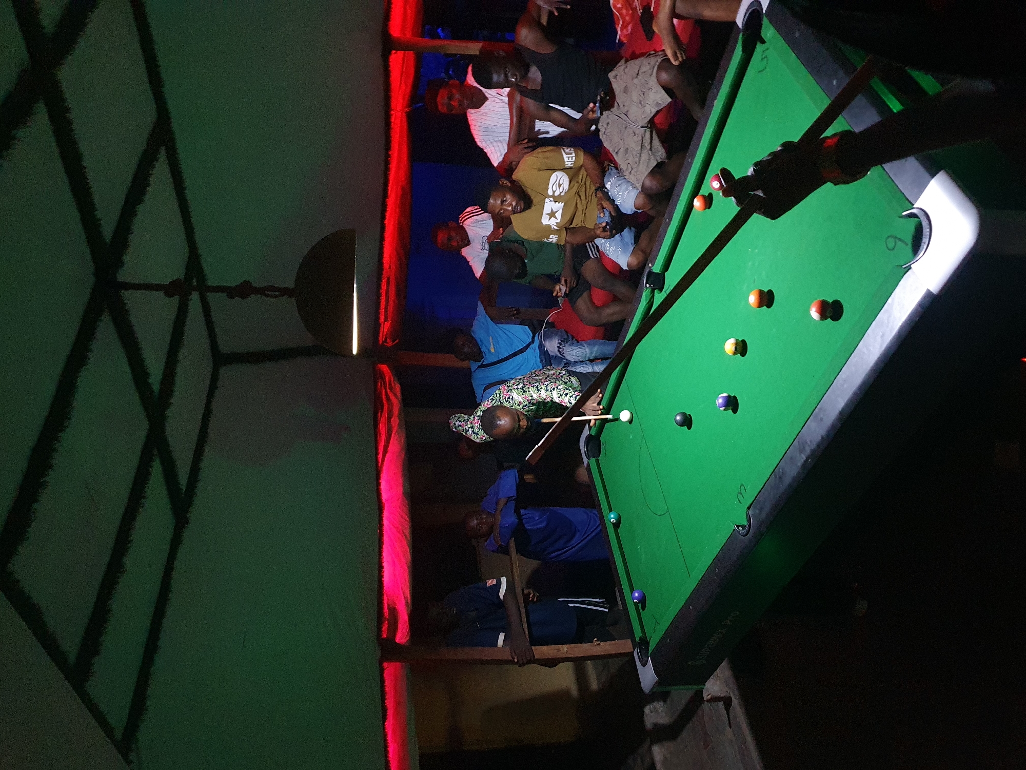 The image depicts an indoor night scene, likely within a pool hall or a casual social venue. The setting is dimly lit, with strong artificial lighting creating contrasting highlights and deep shadows. A vibrant red light source illuminates a vertical surface on the left, while the ceiling above shows a distinct grid-like shadow pattern, possibly from a structural element or decorative screen.
The central focus is a green-felted pool table, upon which several colorful pool balls are scattered. A person, whose hands are visible, is actively taking a shot, holding a pool cue with apparent concentration. Other individuals, mostly men dressed in casual attire, are gathered around the table and in the surrounding areas. They appear to be socializing, watching the game, or simply relaxing, with some standing, others leaning or sitting on benches or low structures. The atmosphere suggests a communal and leisurely evening activity.
Notable details include the stark lighting, which highlights the pool table and players while leaving much of the background in shadow. The text "SUPERMAX PRO" is visible on the side of the pool table, indicating its brand. Faint numbers "6" and "2" can also be seen near pockets on the table. The overall scene captures a moment of social interaction and recreation in a local establishment, consistent with a nighttime setting in Lokoja, Nigeria.