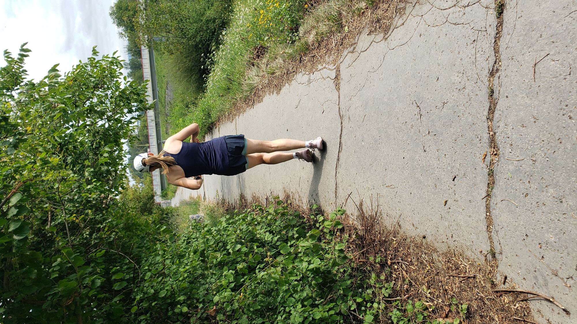 A woman with a ponytail, wearing a dark blue tank top, dark shorts, pink running shoes, and a white baseball cap, runs away from the camera on a cracked asphalt path. The path is bordered on the left by dense green foliage and on the right by dry grass and wildflowers. Overhead, leafy tree branches cast shadows on the path. In the background, a bridge with red railings spans across a body of water or a ditch. The scene appears to be a recreational trail, possibly in the city of Prešov, Slovakia, given the context. The lighting suggests it is daytime, with a partly cloudy sky.