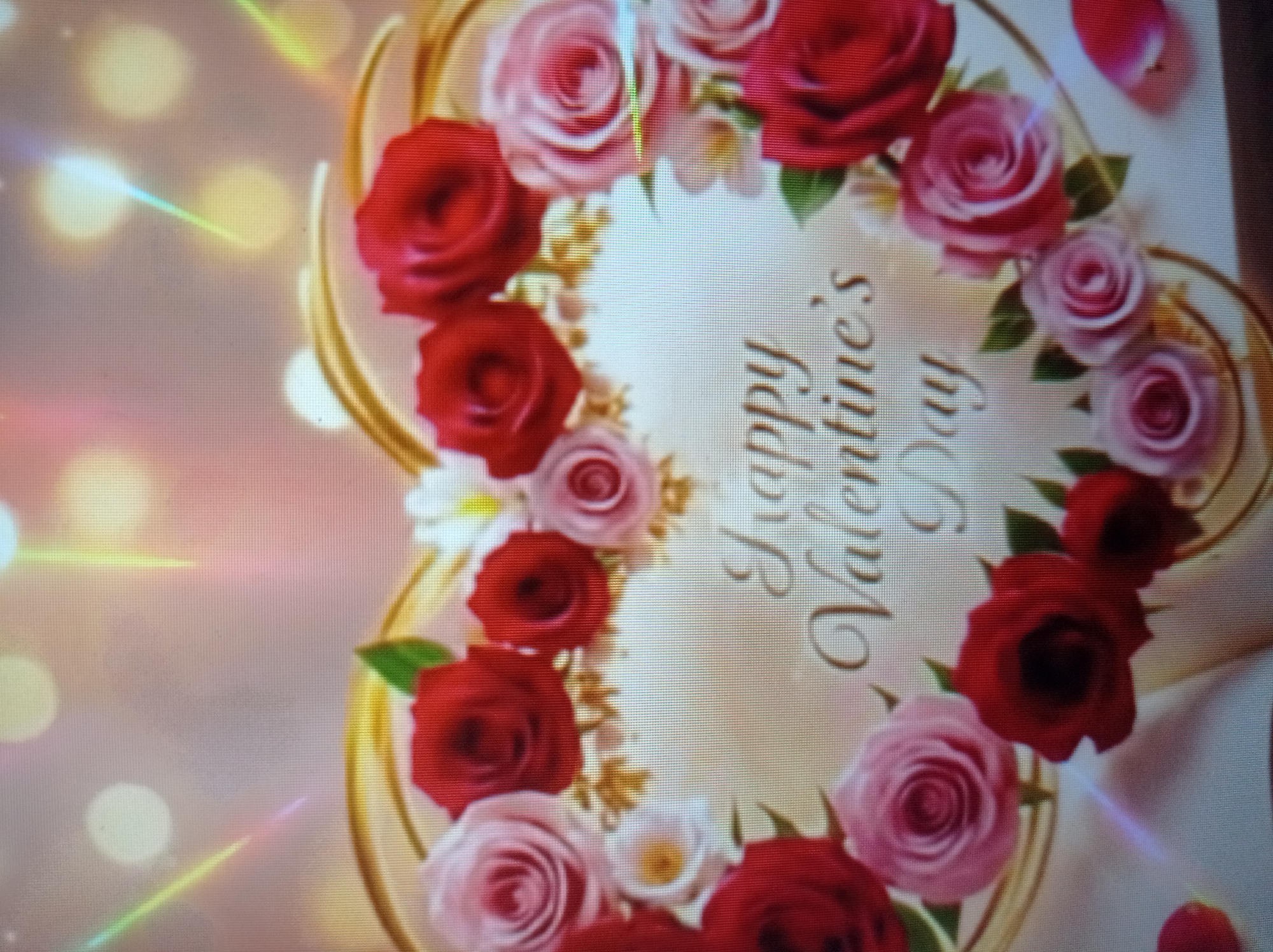 The image is a close-up of a Valentine's Day card. The card is heart-shaped and decorated with an arrangement of red and pink roses, along with smaller white flowers. The words "Happy Valentine's Day" are written in a cursive font in the center of the heart. The background is softly blurred, with bokeh lights and streaks of light that suggest a celebratory or romantic atmosphere. The overall impression is one of a sweet and classic Valentine's Day greeting. There are no visible elements that place this image in Mukachevo, Ukraine, or any specific city.