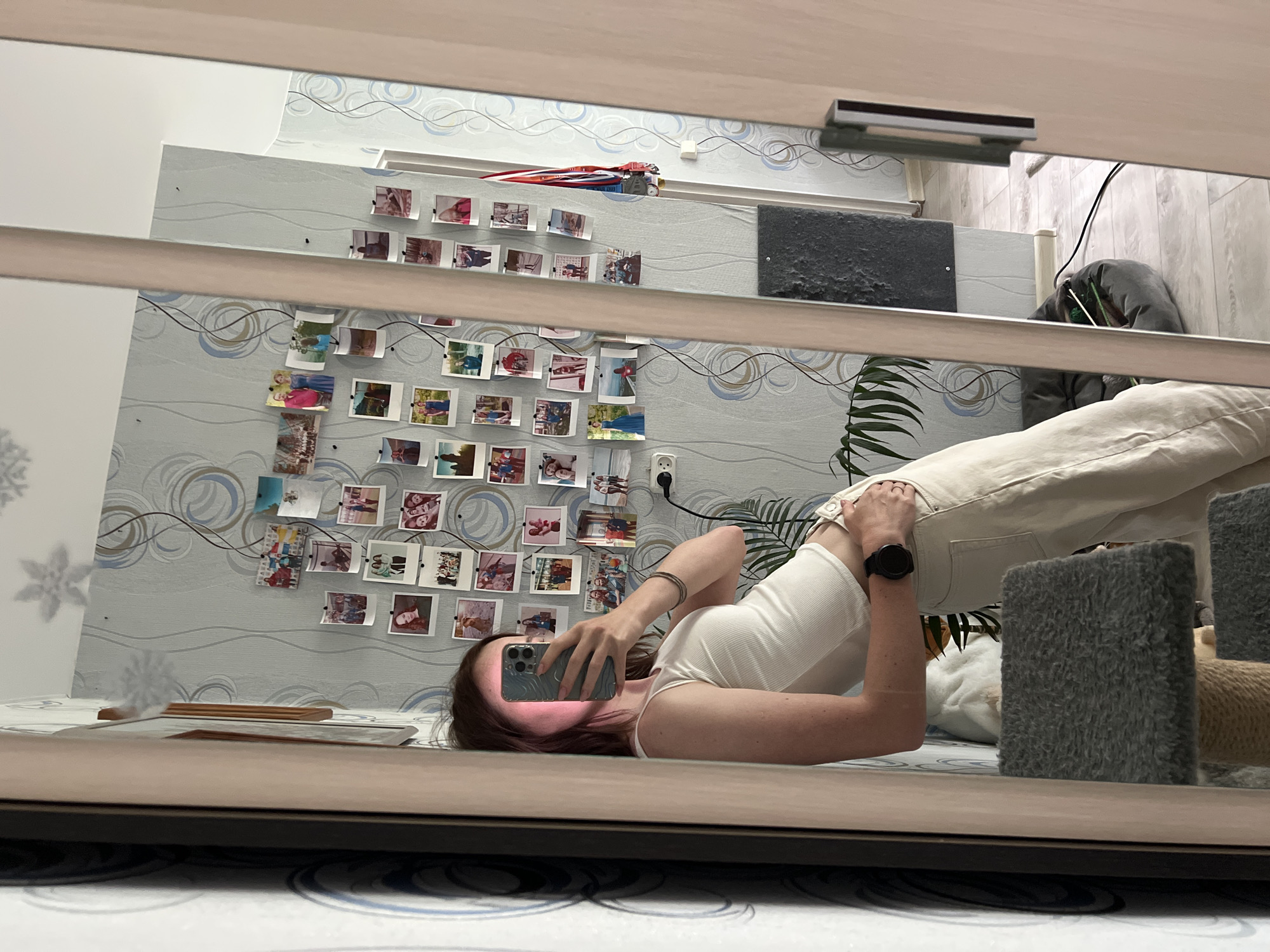 The image shows a person taking a selfie in front of a mirror. The person is lying down, wearing a white tank top and white pants. They are holding a smartphone, presumably to take the picture. The background features a wall decorated with a string of photographs, as well as a plant, and a cat tower. There is a toy car atop a shelf. Based on the visual context (wall covering, general architectural style), there is a likelihood that the image may have been taken in a dwelling within an urban environment. However, there is no specific evidence in the image to associate it with the city of Chaikovsky, Russia.