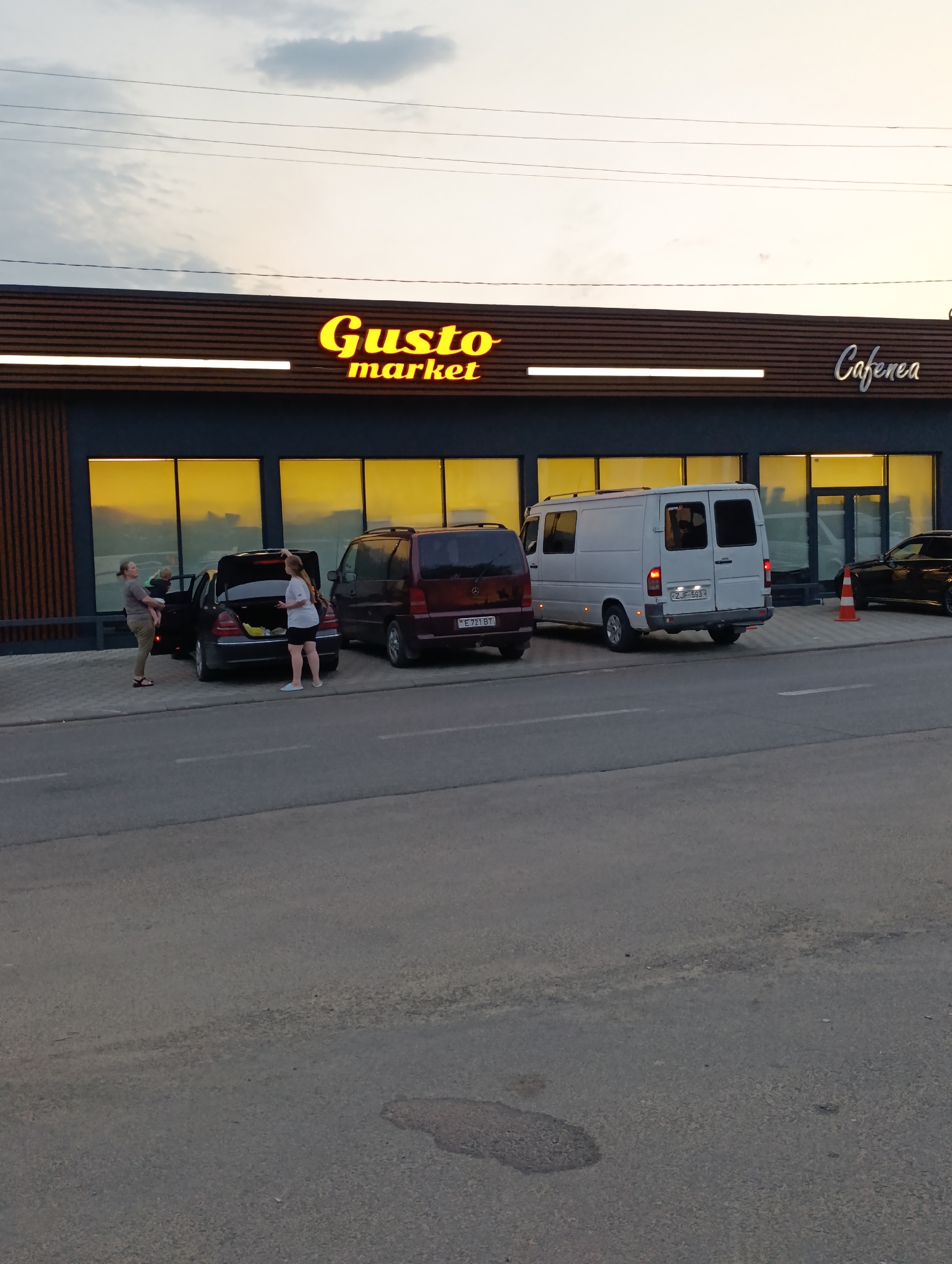 The image captures a scene in front of a building with a prominent "Gusto market" sign in yellow neon letters, and to the right, a sign reads "Cafenea." The building has a dark facade with horizontal wooden accents above large, yellow-tinted windows. In the foreground, a paved road is visible, with a sidewalk area where several vehicles are parked.
On the sidewalk, there is a black sedan with its trunk open. Two individuals are near the car; one woman is holding a child and standing next to the open trunk, while another woman is standing by the passenger side of the vehicle. To the right of the sedan, a dark red van is parked, and further to the right, a white cargo van is also parked. A bright orange traffic cone is positioned on the sidewalk near the white van. A dark colored car is parked further down the sidewalk, partially obscured. The sky above is overcast with soft, diffused light. The overall environment appears to be an outdoor commercial area.