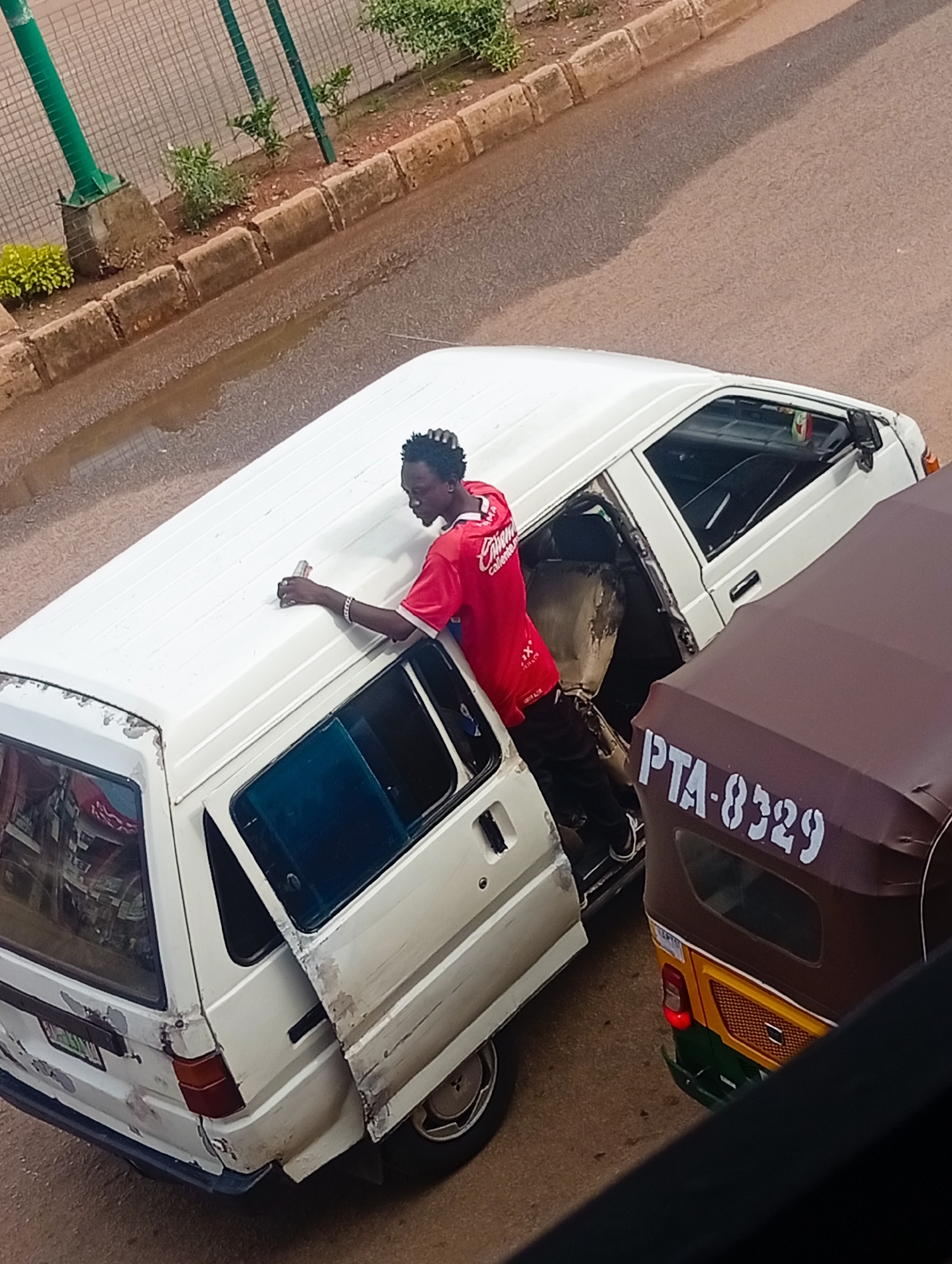 A young man wearing a red shirt and black pants is reaching into the open rear door of a white van. He is holding a can in his right hand. The van appears to be parked on a road in Bukuru, Nigeria, with a brown auto-rickshaw parked next to it. The auto-rickshaw has the license plate "PTA-8329" visible. The road has some puddles of water, and there is a curb with some vegetation along the side. The scene appears to be during daylight hours.