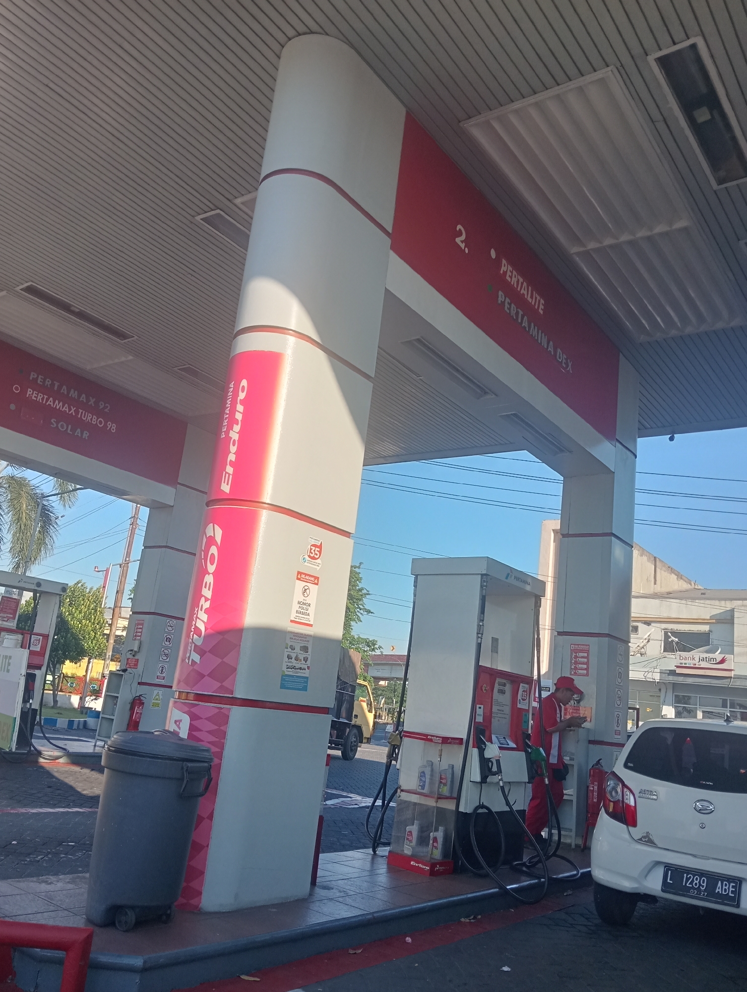 A low-angle shot captures a section of the Pertamina gas station canopy, with a blue sky overhead. In the foreground, a large grey trash receptacle with wheels is positioned on a raised concrete platform. To the right, a white car is parked at a gas pump, with a license plate reading "L 1289 ABE 03-27". A gas station attendant, wearing a red uniform and cap, stands by the pump, engaged in an activity related to the refueling process. To the left, another gas pump is visible, displaying signage for "Pertamax Turbo 92," "Pertamax Turbo 98," and "Solar." The gas station is located in Sawotratap, Indonesia.