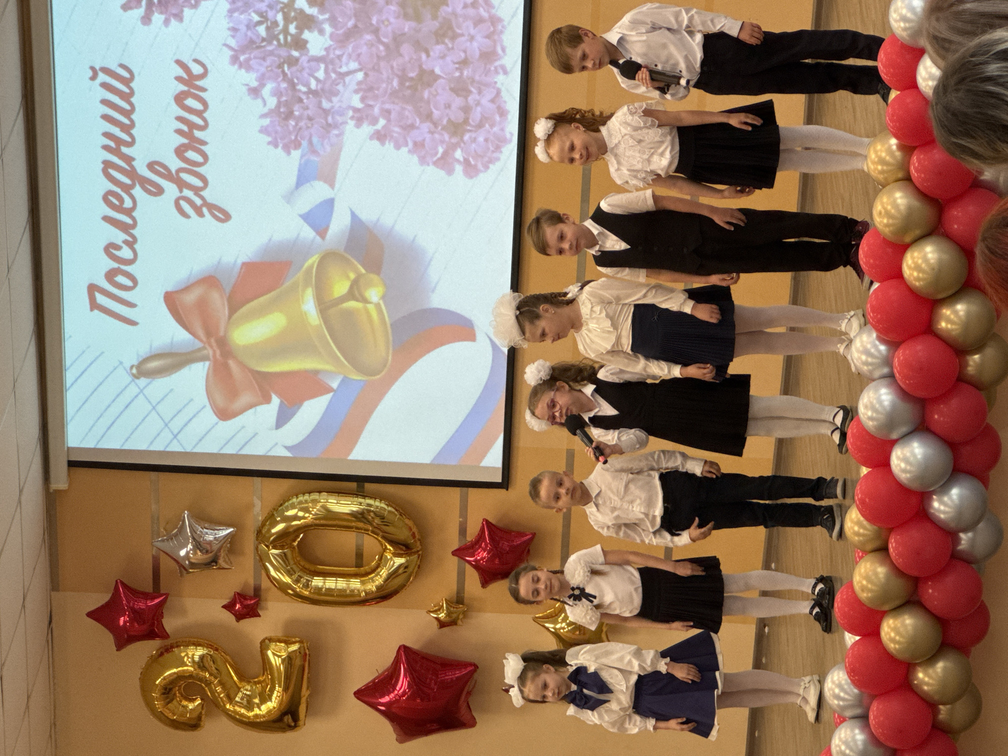 A scene in Ivanovo, Russia, features children on a stage alongside decorations for an event. Seven children, dressed in school attire, including white blouses, dark skirts or pants, and tights, stand side-by-side. One child holds a microphone. Behind the children, a screen displays an image, possibly a slide presentation, with text in Russian and a bell graphic. Balloons in red, gold, and silver adorn the right side of the scene. On the wall to the left, balloon decorations in the shape of stars and the numbers "2020" are visible. The setting is likely a school or community center in Ivanovo.