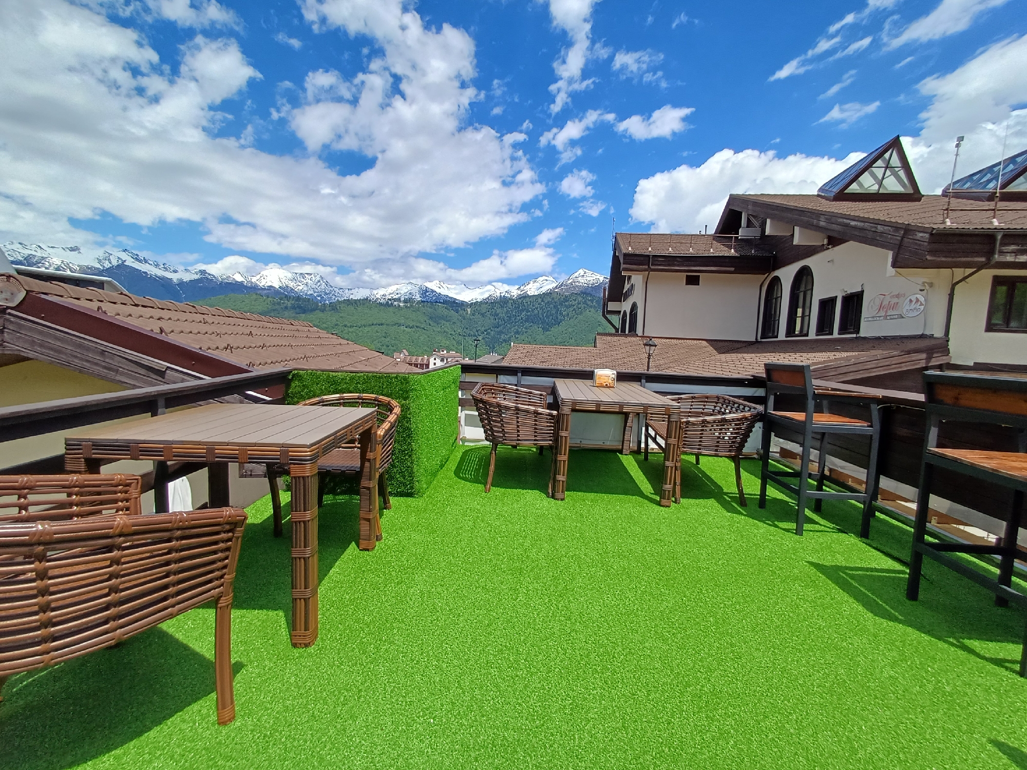 The image captures a bright, sunny daytime scene of an outdoor terrace or patio with a panoramic view of snow-capped mountains. The foreground is dominated by a vibrant green artificial turf floor, creating a stark contrast with the brown and wooden tones of the furniture.
Several sets of outdoor furniture are arranged on the turf. On the left, two low, wicker-style chairs and a rectangular table are visible. In the center, another similar set of two chairs and a table is positioned. To the right, two high, bar-style chairs with dark frames and wooden seats are placed beside a railing. A green artificial hedge acts as a partition on the left edge of the patio. No people are present, indicating a quiet moment.
In the midground, beyond the patio railing, are the rooftops of several buildings with brown tiled roofs. One prominent building features white walls, dark arched windows, and brown trim, with two distinct triangular skylights on its roof. A sign on this white building displays text, which appears to read "TefuR", "БИСТРО" (BISTRO), and "ГОРЫ" (GORY - Mountains).
The background showcases a majestic range of snow-capped mountains under a clear blue sky dotted with scattered white clouds. The lower slopes of the mountains are covered with dense green forests. The weather is bright and clear, with strong sunlight casting distinct shadows from the furniture. The overall scene suggests a high-altitude resort or vacation area, possibly in springtime or early summer given the snow on the peaks and green foliage below.