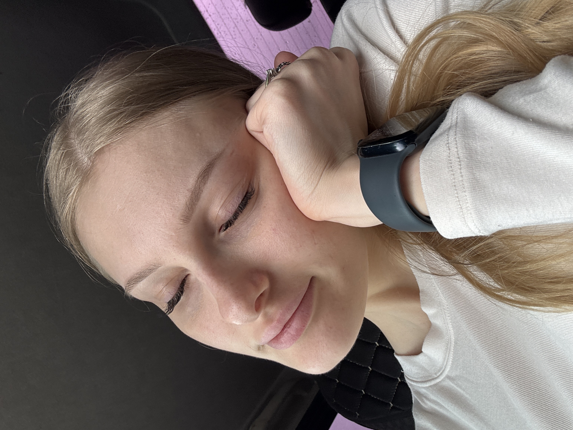 A woman with light blonde hair is lying down, resting her face on her hand. She has her eyes closed and appears relaxed. She is wearing a light-colored shirt. Her wrist has a smartwatch on it. The background is a mixture of black and pink. There is no evidence in the image to suggest its location within Perm, Russia.