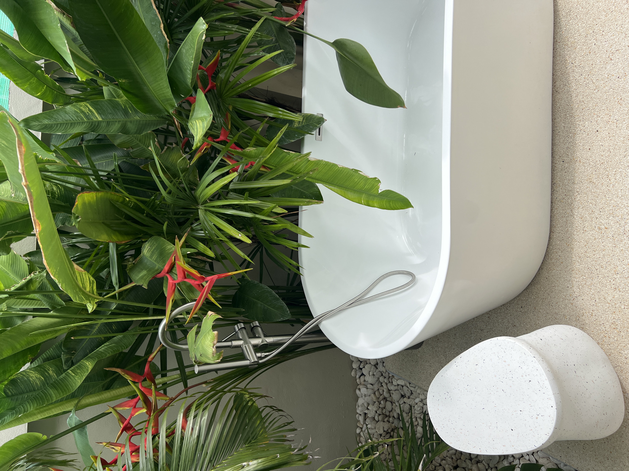 This image depicts a freestanding white bathtub situated amidst lush greenery. The surrounding foliage includes various plants with large green leaves and striking red flowers. A silver faucet and shower hose are visible next to the tub. A two-tiered, speckled white stool rests on a pebbled surface adjacent to the tub. The environment suggests an outdoor or partially outdoor bathroom setting. The location is unspecified.