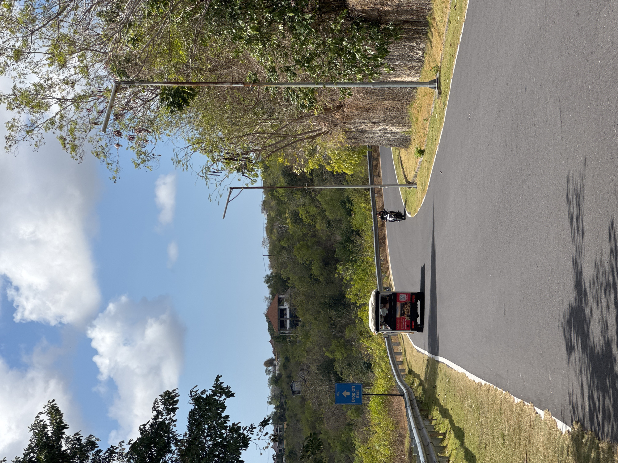 The image depicts a sun-drenched, curved asphalt road in Kuta Selatan, Indonesia. Lush green vegetation covers the rolling hills in the background, interspersed with a few buildings. The sky is a vibrant blue, adorned with fluffy white clouds. On the right side of the road, a steep, rocky embankment is visible, lined with trees and vegetation. A modern street lamp stands tall beside it.
Two vehicles are present on the road: a small, red and white electric vehicle with a canopy, resembling a golf cart, is moving away from the viewer on the left side of the road, and a lone motorcyclist is visible further up the road on the left. A blue sign with a white arrow indicating "Drop Off Exit" is placed on the left side of the road, near the electric vehicle. The road is bordered by a metal guardrail on the left and a grassy shoulder on the right. Shadows of trees are cast across the road, indicating it is daytime.