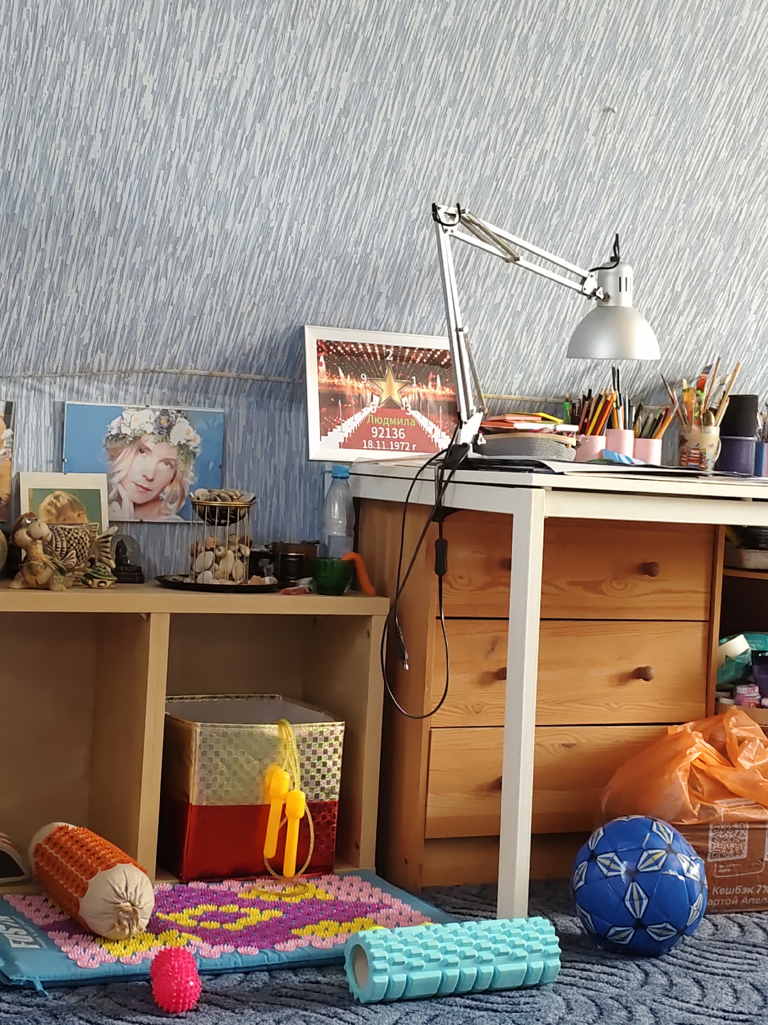The image depicts an indoor scene, likely a desk area in a home or studio. The background is a blue and white textured wallpaper. On a wooden shelf, there are several framed pictures, a turtle-shaped figurine, and a decorative holder filled with pebbles. A desk with wooden drawers is visible, equipped with a metal desk lamp and an assortment of art supplies, including pencils and paintbrushes in containers. To the right of the desk, an orange plastic bag and a cardboard box are present, along with a blue soccer ball on the carpeted floor. In the foreground, a blue yoga mat with a colorful pattern is laid out, featuring a corn-shaped item, a pink spiked ball, and a blue foam roller. The overall impression is of a creative or hobby-focused space. There is no discernible connection to the city of Rostov-on-Don, Russia.