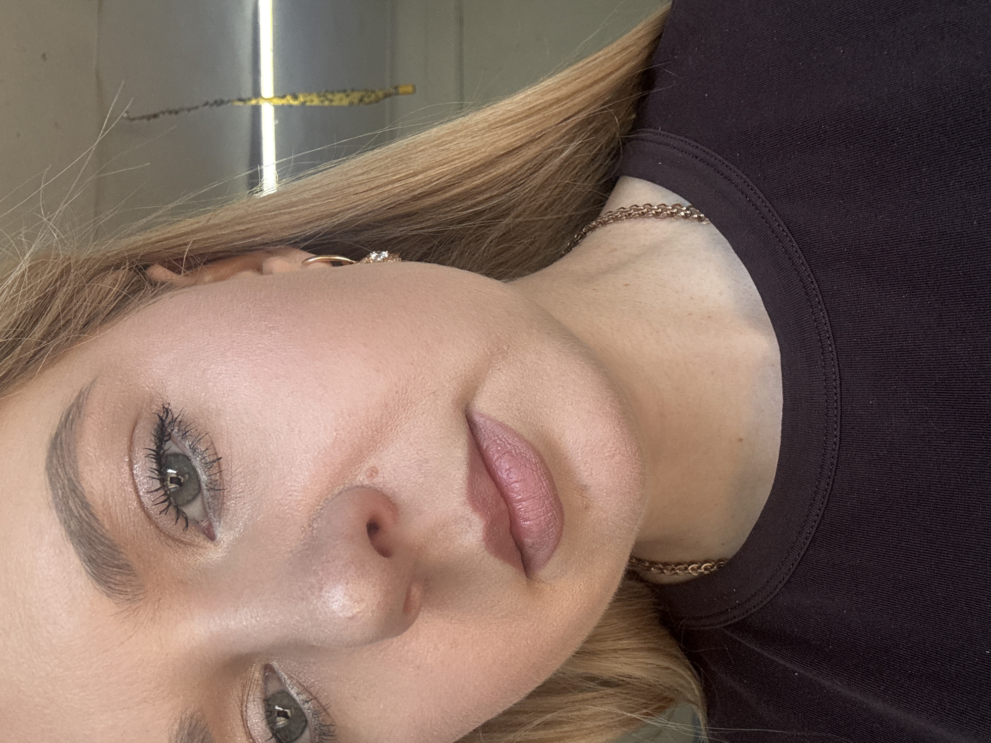 A close-up, portrait-style, outdoor, eye-level, slightly high-angle, natural-light, medium-shot photograph shows the right side of a young adult Caucasian woman's face. The woman has fair skin, long blonde hair, and light green eyes. Her eyebrows are dark and arched, and she is wearing light makeup, including mascara and eyeshadow. She also has a small stud earring in her right ear. She is wearing a dark t-shirt and a gold chain necklace. The background is out of focus and appears to be a concrete wall with some yellow and black striped caution tape hanging from above. The lighting suggests it is daytime, and the overall tone is neutral.