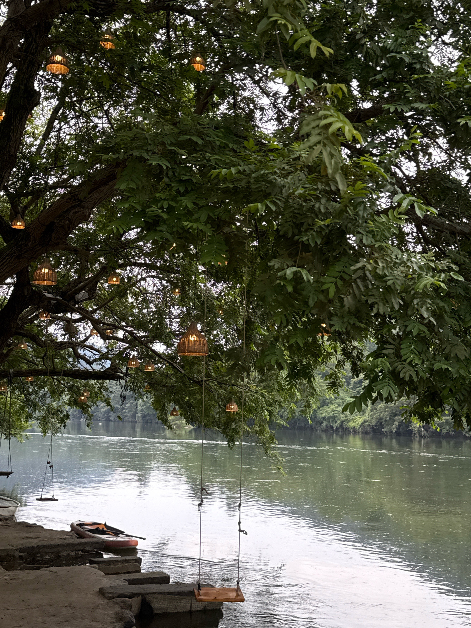 The image depicts a serene riverside scene, likely in Yiwu, China, characterized by lush greenery and tranquil waters. A large tree with dense foliage overhangs the bank, adorned with numerous woven lantern-style lights that cast a warm glow. Hanging from the tree branches are several swings, some with wooden seats and others with rope seats, suggesting a recreational area.
On the left side of the frame, a stone embankment leads down to the water. Resting on the edge of the embankment, partially in the water, is an inflatable paddleboard with a paddle. To the left of the paddleboard, a white canoe or kayak is also visible. The river itself is calm and reflects the overcast sky and surrounding greenery. The opposite bank of the river is lined with dense trees and vegetation, creating a natural, unspoiled backdrop.
The overall atmosphere is peaceful and inviting, hinting at a quiet spot for relaxation and outdoor activities. The presence of the swings and watercraft suggests this location is utilized for leisure by individuals or groups. The lighting, even in daylight, adds a touch of ambiance. The sky is overcast, suggesting a soft, diffused light. No people are visible in the image.