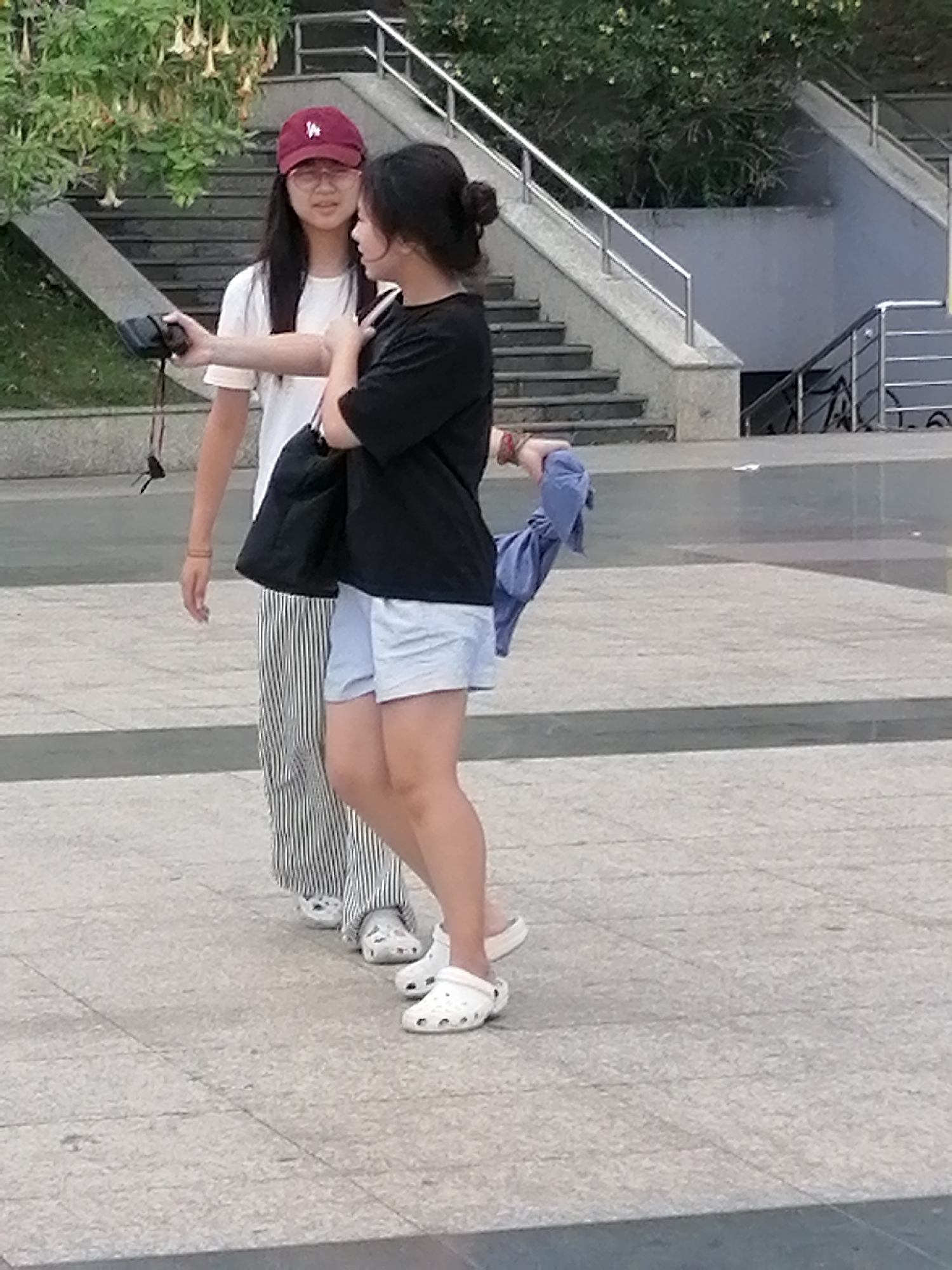 The image shows two young women standing on a paved plaza. The woman on the left, wearing a red baseball cap and a white t-shirt with black and white striped pants, holds a black camera with a strap. The woman on the right wears a black t-shirt and light blue shorts, holding a crumpled blue garment in her right hand. Both women are wearing white Crocs. In the background, there are stairs with metal railings, greenery, and some hanging yellow flowers. The scene is likely outdoors, possibly in a public area of Da Lat, Vietnam, given the presence of stairs and paved ground common in urban settings.