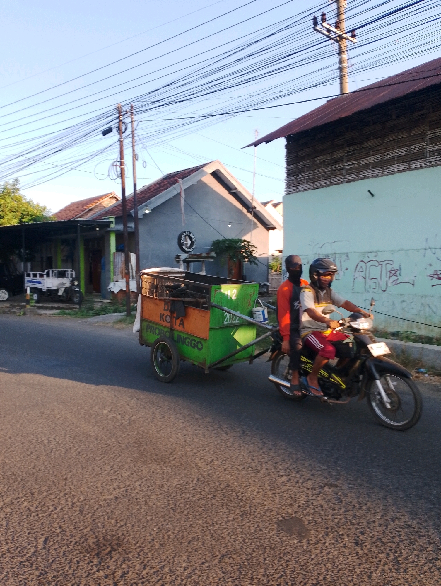 The image depicts a scene on a street in Probolinggo, Indonesia, with a motorcycle carrying two passengers and pulling a trailer. The motorcycle is black with yellow accents and has a license plate that is partially obscured. The driver, wearing a helmet and a mask, is dressed in a light-colored shirt with dark stripes on the collar and arms, and red and white striped shorts. The passenger behind the driver is wearing an orange long-sleeved shirt and a dark head covering, obscuring their face. The trailer is green and orange, with writing on its side that reads "KOTA PROBOLINGGO 2022". In the background, there are several buildings, including houses with tiled roofs and a building with a light blue facade and wooden siding. Electrical wires are strung across the sky, supported by utility poles. Trees and other foliage are visible on the left side of the image, and the road surface is asphalt. The lighting suggests it is either early morning or late afternoon, with sunlight casting shadows.