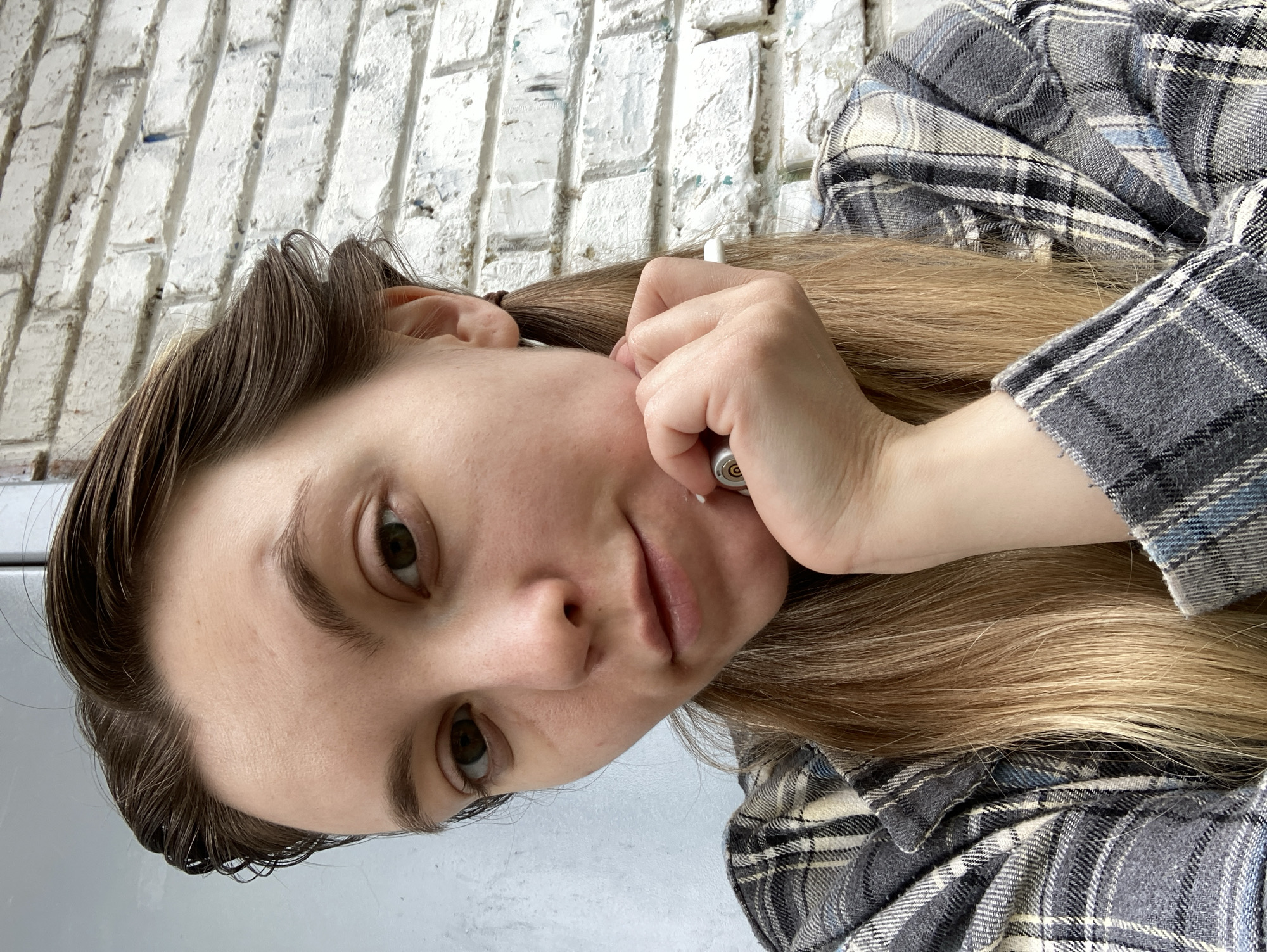 A close-up selfie of a woman with shoulder-length blonde hair, wearing a grey and blue plaid shirt. She is holding a small, cylindrical object near her face, possibly a pen or cosmetic tool. Her expression is neutral. In the background is a white brick wall. The location of Chaikovsky, Russia cannot be verified from this image alone.