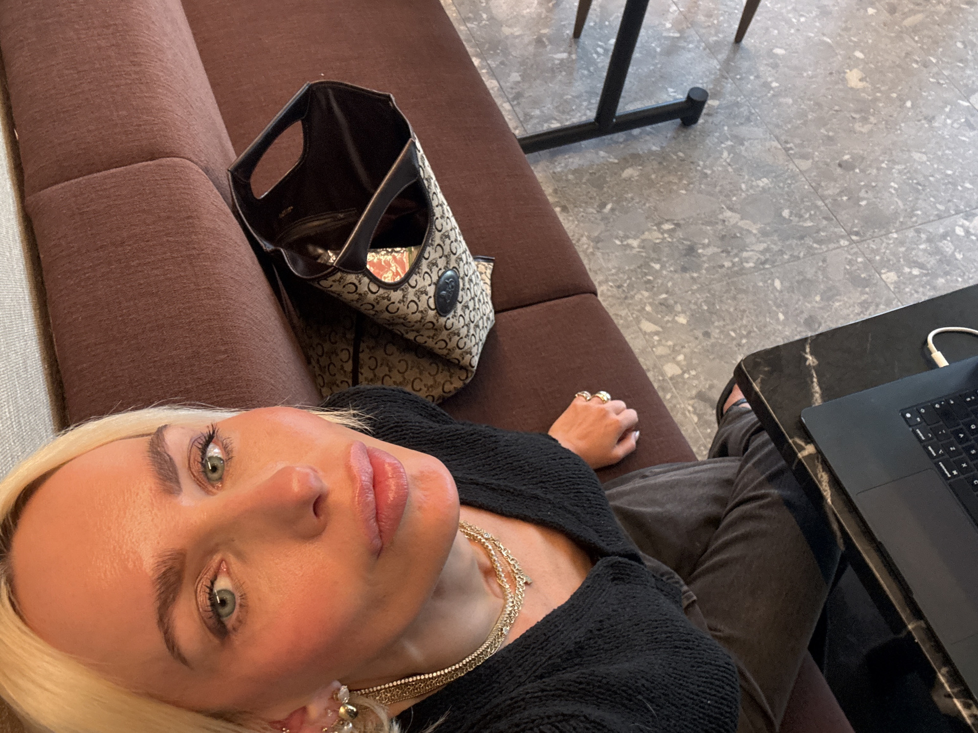 An overhead, wide-angle selfie captures a blonde woman relaxing indoors, likely in San Diego, United States of America. The woman, with fair skin and light green eyes, wears visible makeup including mascara, eyeliner, and lip gloss. Her light hair is pulled back, and she is adorned with multiple gold necklaces and an ornate earring. Rings are visible on her right hand. She wears a black, textured long-sleeved top and dark pants, visible down to her legs.
She is reclining on a brown, upholstered sofa or bench. Next to her, on the sofa, sits an open handbag featuring a repeating dark brown C-like or horseshoe-like monogram pattern against a lighter background.
To the right of the woman and the sofa, a dark-colored table, possibly marble-topped, holds a partially visible black laptop, likely a MacBook, with its keyboard and trackpad visible. The floor is composed of light-colored, patterned tiles, possibly terrazzo, with dark grout lines. In the background, part of a black metal chair or table leg is visible. The overall setting appears to be a modern lounge, cafe, or waiting area. The lighting suggests it is daytime. No discernible text is clearly legible apart from the repeating pattern on the handbag.