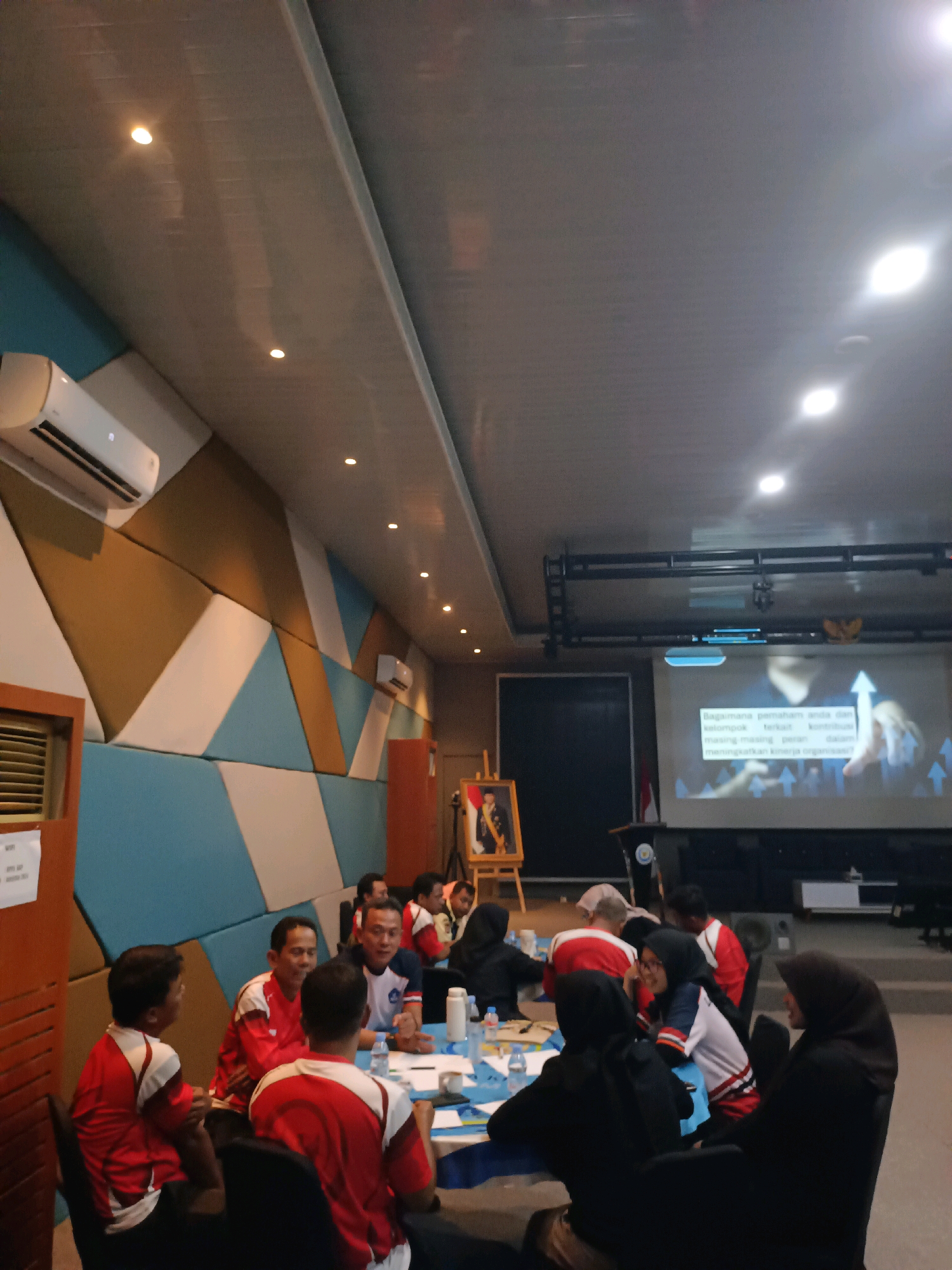 In a meeting room, several people are seated around tables. A projection screen displays an image with text in Indonesian, showing upward trending arrows. Some individuals are wearing red and white uniforms. The room has a modern design with geometric wall panels and track lighting on the ceiling. An air conditioning unit is visible on the wall. The overall impression is that of a training session or a conference in Subang, Indonesia.
