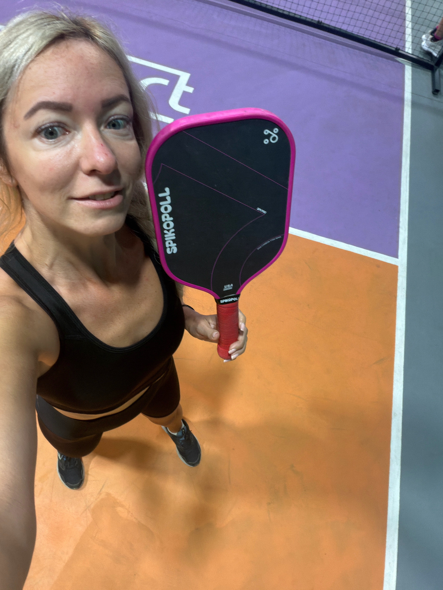 A blonde woman with blue eyes and fair skin, wearing a black sports bra and black shorts, is holding a pink and black pickleball paddle. She is standing on a pickleball court with purple and orange surfaces, and a grey area with a white line. The court has a net in the background. The woman is looking at the camera and smiling. She has on dark athletic shoes. The lighting suggests an indoor environment. The image does not provide any information or context that would place it in Da Lat, Vietnam.