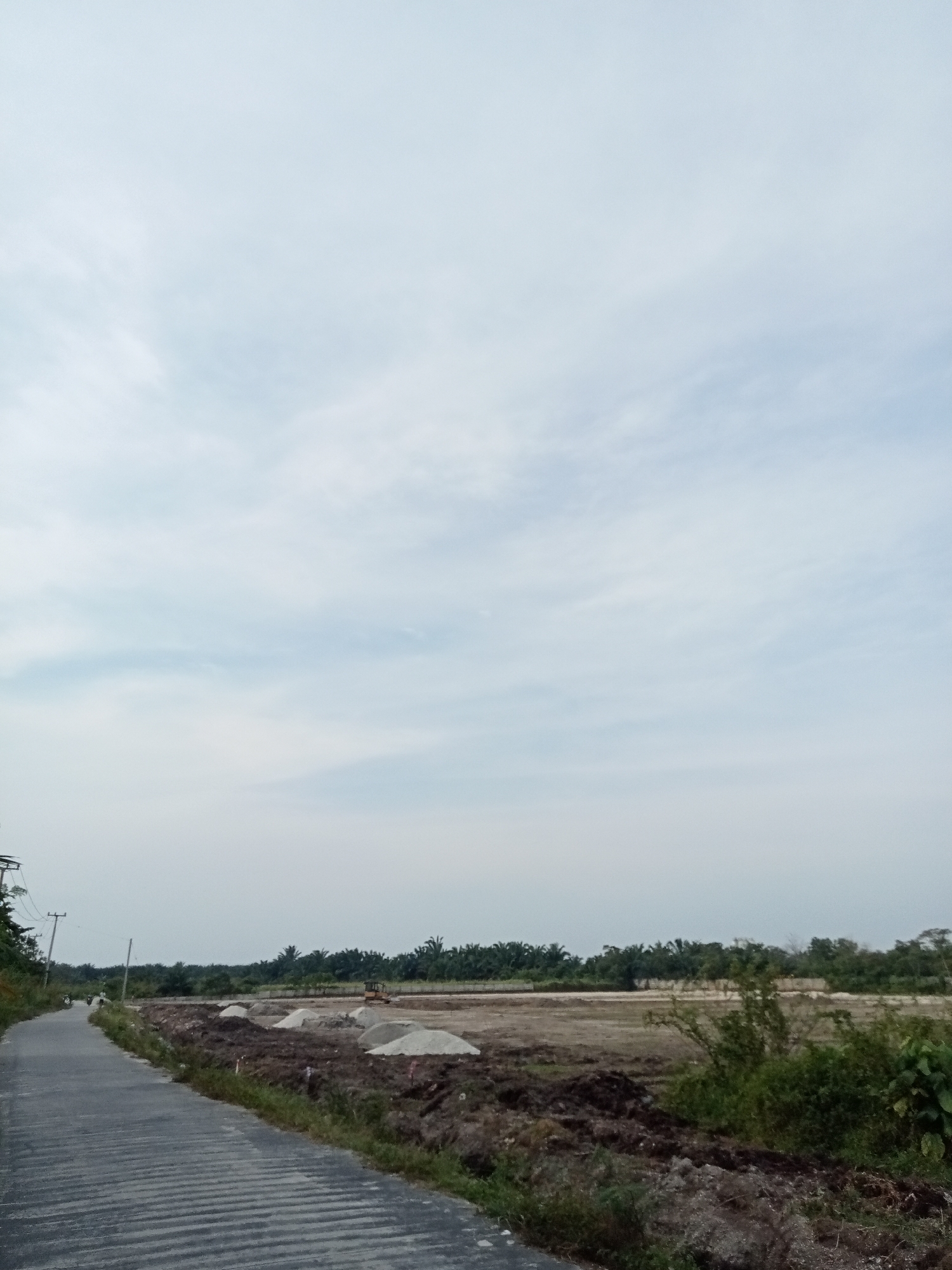 The image depicts an overcast sky above a rural scene in Teluk Kenidai, Indonesia. In the foreground, a paved road with distinct horizontal ribbing runs diagonally from the lower left to the mid-left of the frame. To the right of the road is an area of disturbed earth, with mounds of sand and piles of dark soil. A small yellow excavator is visible in the mid-ground on the construction site. A line of palm trees forms a distant treeline, separating the construction area from the sky. A power pole with wires is situated on the left side of the road, and two motorcycles are seen on the road in the distance. The overall environment appears to be a developing area, possibly undergoing construction or land preparation.