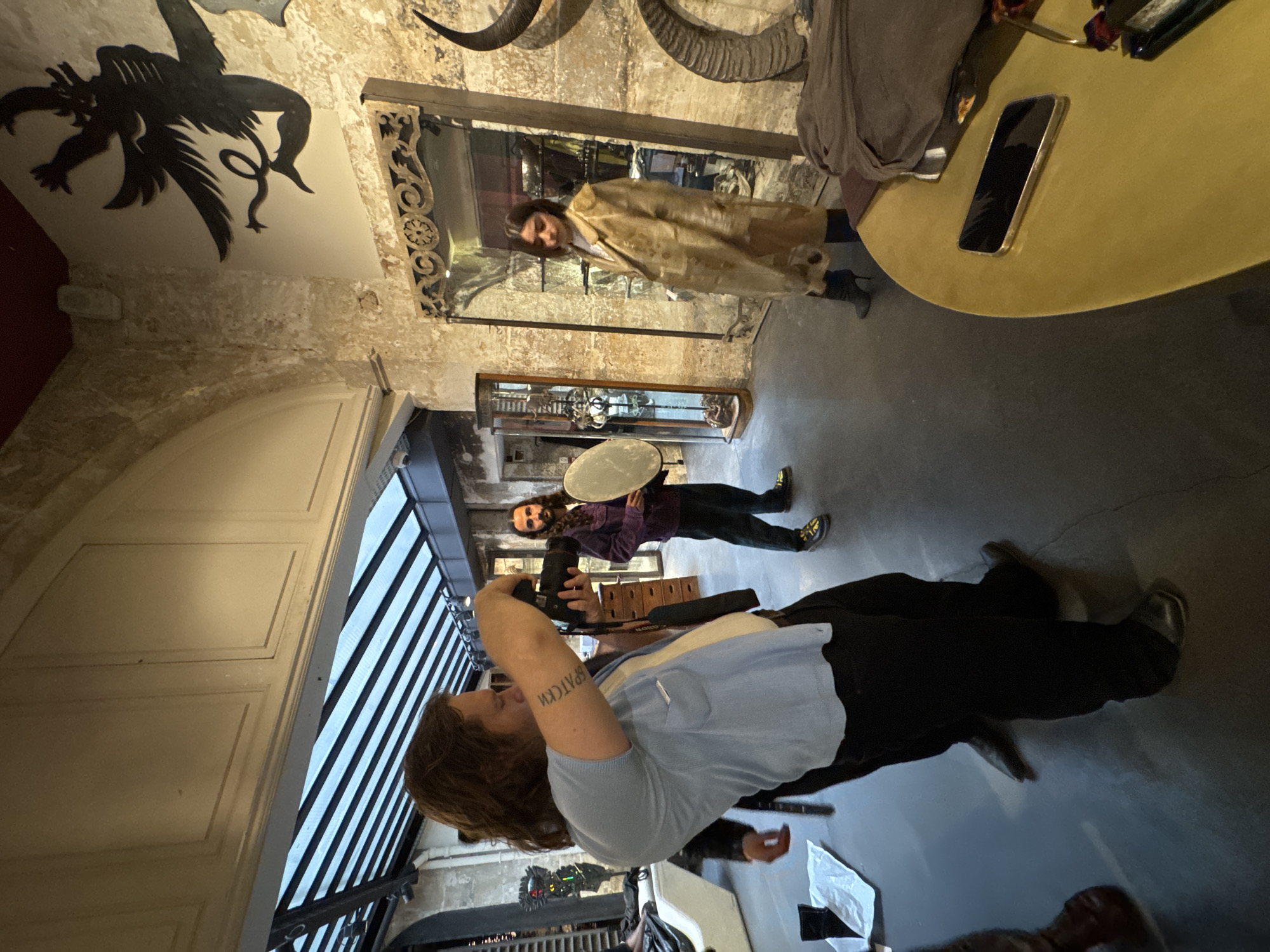 The photo depicts an interior scene with several people present. The dominant figure in the foreground is a person holding a camera, presumably taking a photograph. A second individual stands further back, holding a reflector. A third person is visible in the background, reflected in a mirror.
The setting appears to be a retail space, given the displays and objects scattered around, and the architecture suggests a historic building. The presence of taxidermied animal horns mounted on the wall adds a distinctive element to the decor.
While I cannot confirm the location within Paris, France with certainty, the architectural style and interior design suggest a store or shop with a unique aesthetic, potentially situated in an older part of the city.