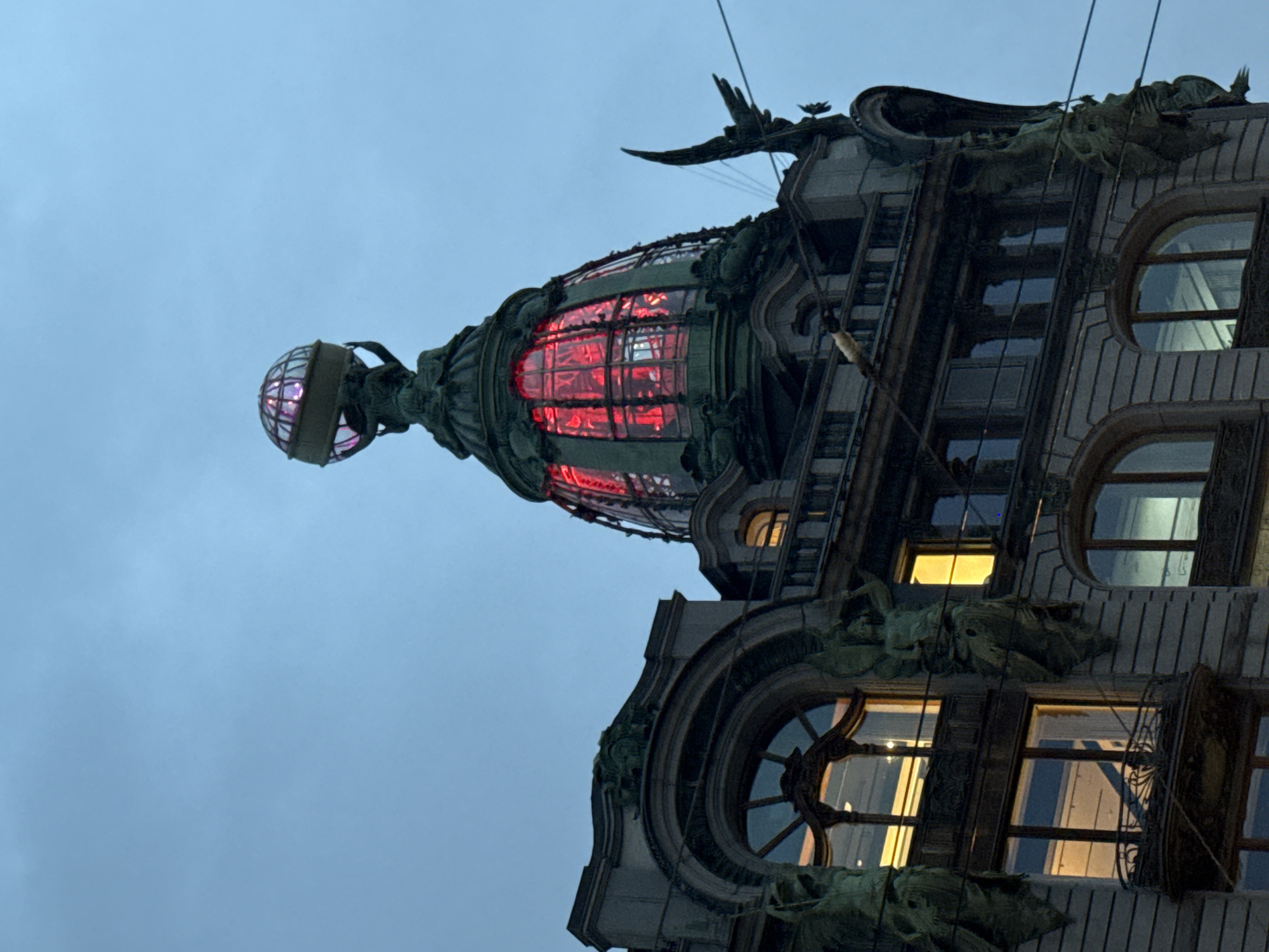 The image shows the Singer House (Dom Knigi) in Saint Petersburg, Russia, a historic building located on Nevsky Prospekt. The perspective is from below, looking up at the upper floors and the iconic globe-topped dome. The sky is a muted blue, suggesting twilight or an overcast day.
The building's facade is ornate, with classical architectural elements and decorative sculptures. On the left side, figures resembling winged atlantes or caryatids are integrated into the stonework, holding up architectural features. Similar sculptures are visible on the right side of the building. The windows on the lower floors are illuminated from within, casting a warm yellow light. Some of these windows are arched, and one features a small balcony with decorative metalwork.
The prominent feature is the green copper dome, which is elaborately decorated and has stained-glass panels illuminated with a vibrant red light. At the very top of the dome sits a large, metallic globe held aloft by a sculpted figure. To the right of the dome, a black, winged sculpture, possibly representing an eagle or another mythical creature, is perched on the building's corner. Several power lines are visible crossing the frame, adding a touch of urban reality to the grand architecture. The overall impression is one of grandeur and architectural detail against a somber sky.
