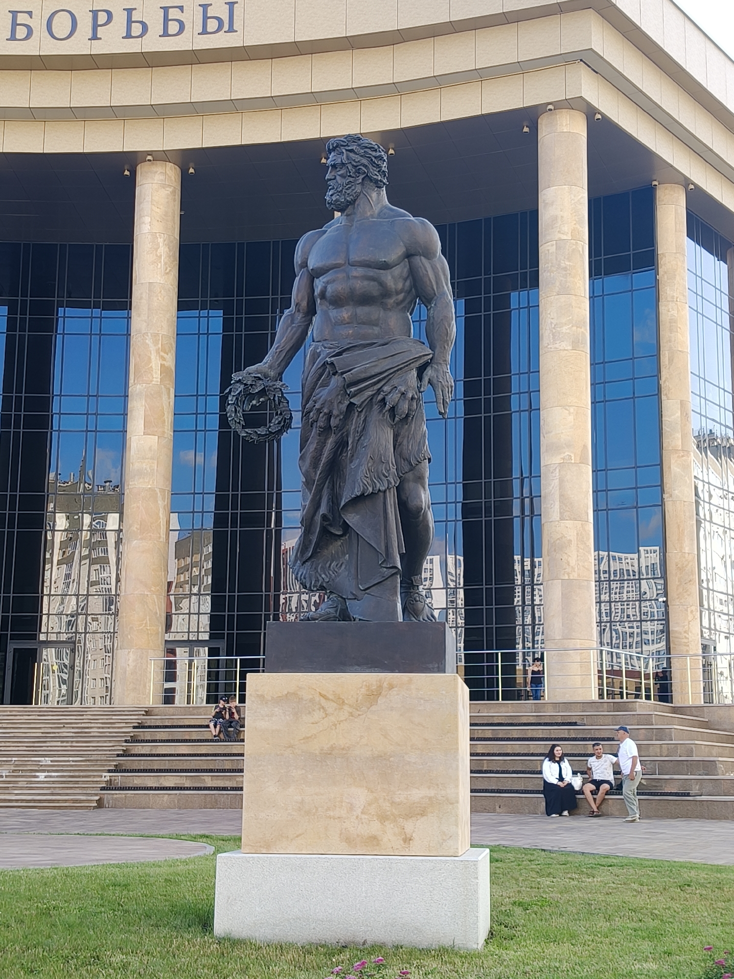 The image depicts a large bronze statue of a muscular man holding a wreath in front of a modern building with a glass facade. The building's facade is highly reflective, showing the surrounding environment. The statue stands on a stone pedestal. In the foreground, there is a well-maintained lawn. Further back, a set of stairs leads up to the building. Several people are visible on the stairs and in front of the building, some sitting and others standing. The sky appears to be clear. The overall scene suggests an urban public space, possibly a monument or a landmark.