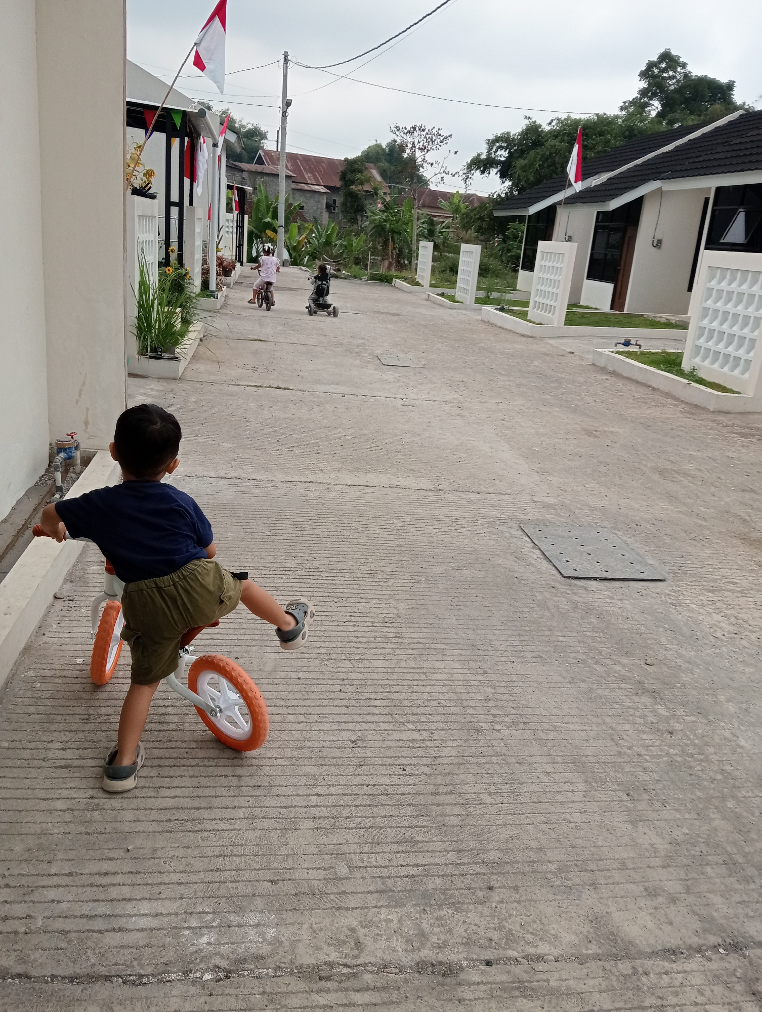 In Candirejo, Indonesia, on a cloudy day, a young boy wearing a dark blue t-shirt and olive green shorts is riding a white balance bike with orange wheels. He is on a paved road in front of a row of modern, light-colored houses with dark roofs. The houses are decorated with Indonesian flags and pennants. In the background, two other children are visible on smaller bicycles or tricycles, further down the road. Trees and vegetation are present behind the houses. The road has a concrete surface with visible parallel grooves. A metal grate is set into the road to the right of the boy. The scene depicts children playing in a residential neighborhood.