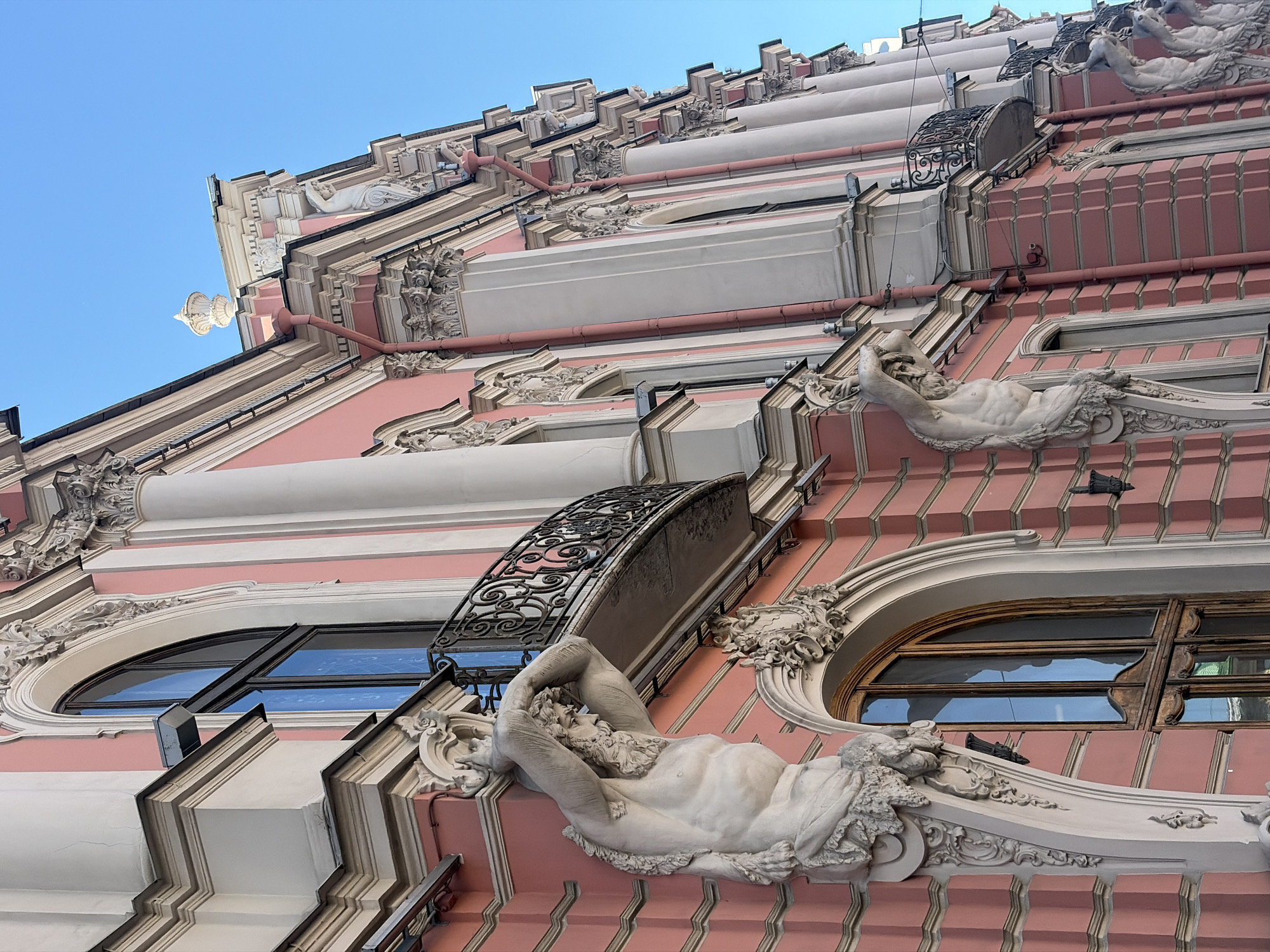 The image shows a close-up, low-angle shot of a pink Baroque building in Saint Petersburg, Russia. The facade features prominent white Corinthian columns, arched windows with dark frames and wooden sills, and ornate architectural details. Two large, muscular statues of male figures, possibly atlantes or caryatids, support the balconies above, their bodies carved in stone and detailed with beards and flowing hair. The balconies themselves have black wrought-iron railings. The sky is a clear, bright blue. The building's pink stucco walls are textured and have horizontal banding. The architecture suggests a grand, historical structure, likely a palace or a prominent public building in the city.