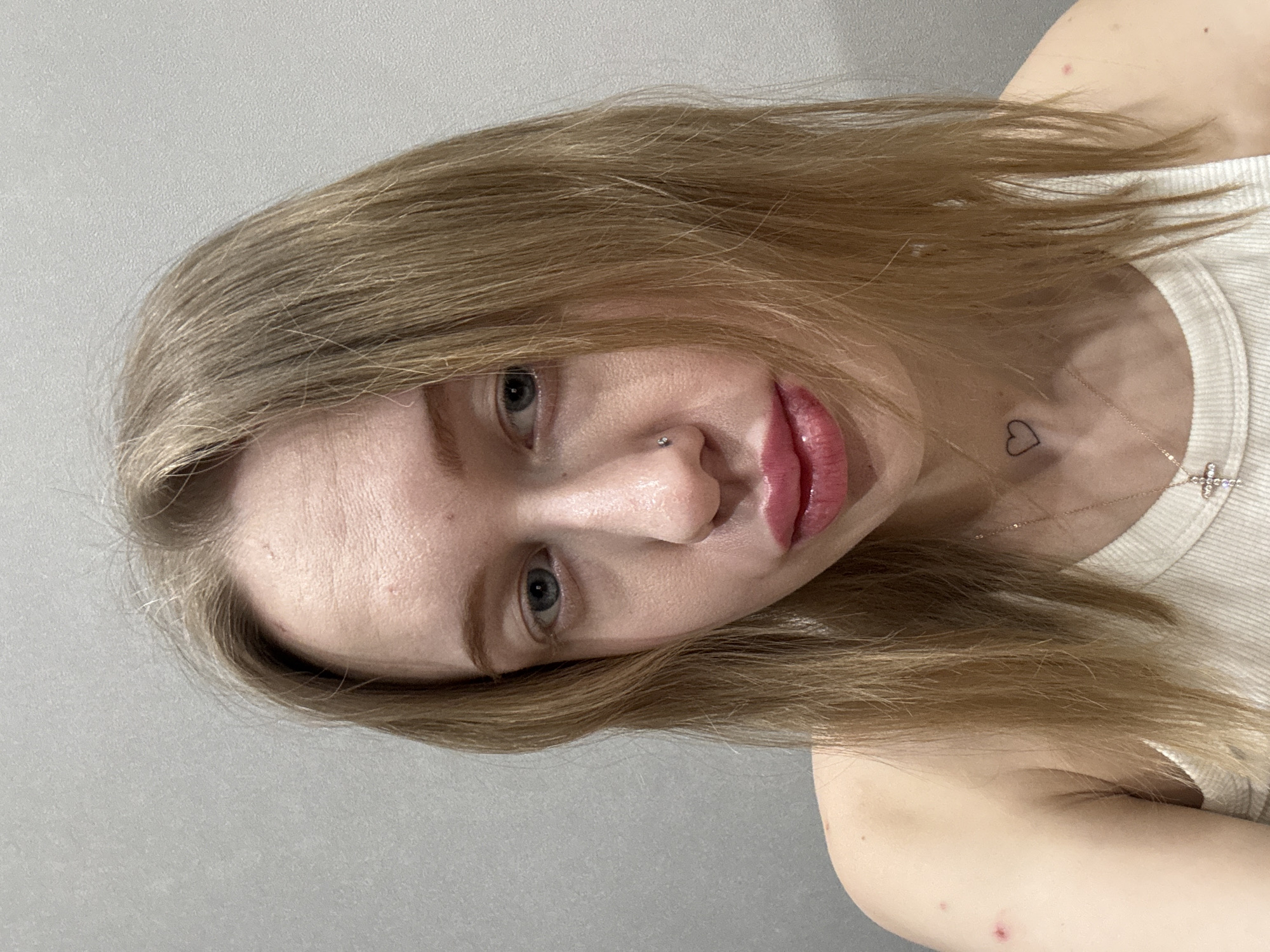 A young adult female is depicted in a close-up, frontal portrait, gazing directly at the viewer. She has blonde hair, blue eyes, and a fair complexion. A small, silver-colored stud is present in her left nostril. Her lips are full, appearing moisturized or glossed with a pink hue. A small, black outline heart tattoo is visible on the left side of her neck. She wears a white ribbed tank top and a thin, gold-toned necklace with a small, clear-stoned cross pendant. Minor skin imperfections are discernible on her forehead and both shoulders. The background consists of a plain, light grey, textured wall, indicating an indoor setting. The scene is located in Rostov-on-Don, Russia; the precise interior location within the city is not visually identifiable from the image.