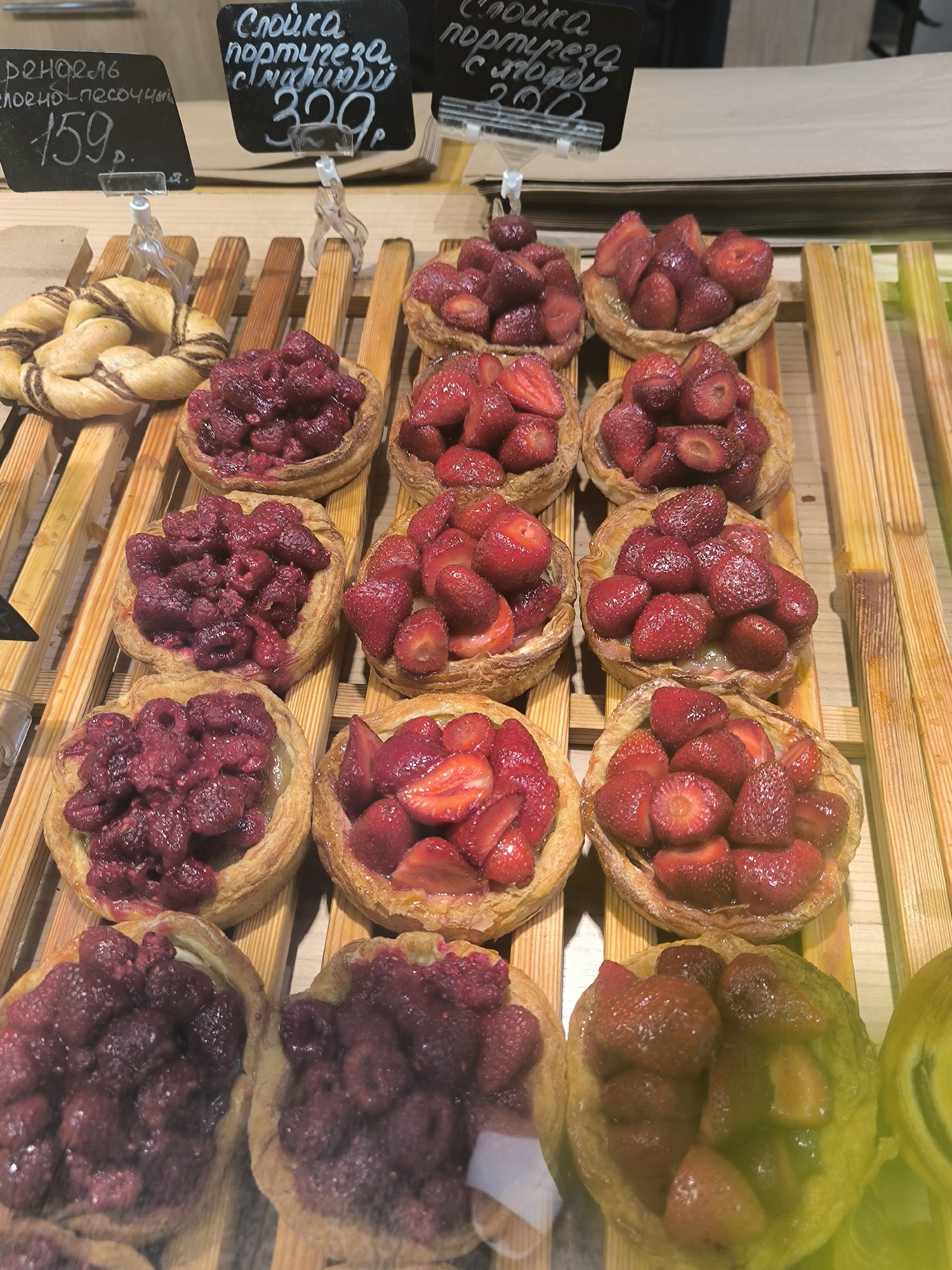 A close-up, eye-level shot displays a variety of freshly baked pastries arranged on wooden shelving in what appears to be a bakery or market stall in Novosibirsk, Russia. The pastries are small tarts with flaky, golden-brown crusts filled with berries. From top to bottom, and left to right, the visible pastries include: a twisted, pretzel-like bread with chocolate filling; several tarts filled with raspberries; multiple tarts filled with sliced strawberries; and in the foreground, two tarts filled with raspberries and one tart filled with a mix of strawberries and what appears to be kiwi or green apple slices. Black chalkboards with handwritten Russian text and prices are visible above the pastries, indicating product names and costs. The lighting is bright, highlighting the textures and vibrant colors of the fruits and baked goods. No people are visible in the frame, but the scene suggests a commercial establishment. The overall impression is one of fresh, artisanal baked goods ready for sale.