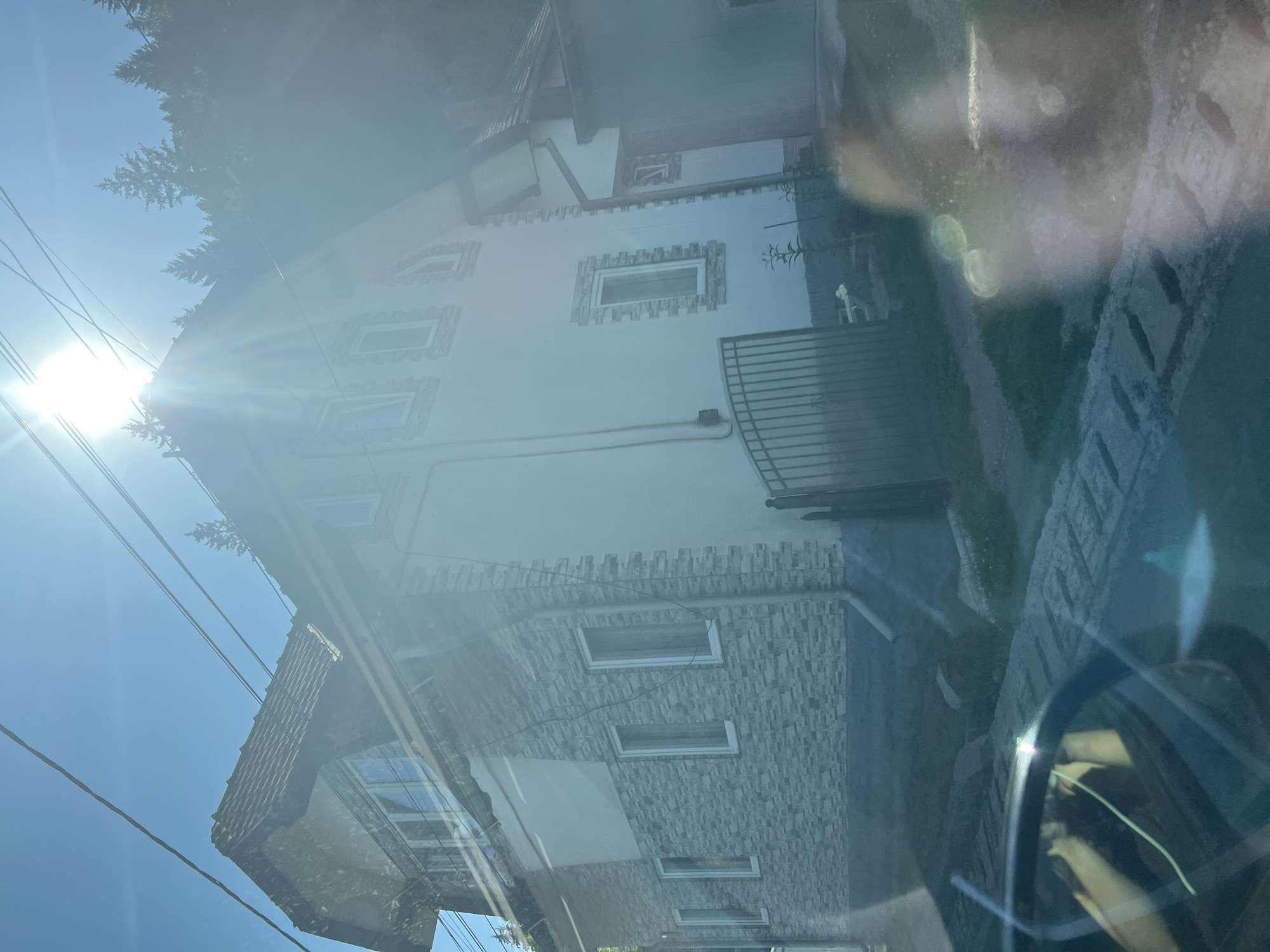 This is a photograph taken from inside a vehicle, looking out at a house in Prisaca Dornei, Romania. The sun is bright and causes lens flare in the image. The house appears to be a multi-story building with a sloped roof covered in tiles. The exterior walls are a mix of white plaster and grey stone-like siding. There are several windows visible on the house, some rectangular and some arched. A metal fence is located in front of the house. Power lines are visible strung across the sky. The environment appears to be a rural or suburban setting, with trees and vegetation visible in the background. The foreground shows part of the vehicle's interior, including the dashboard and windshield, with reflections and glare present.