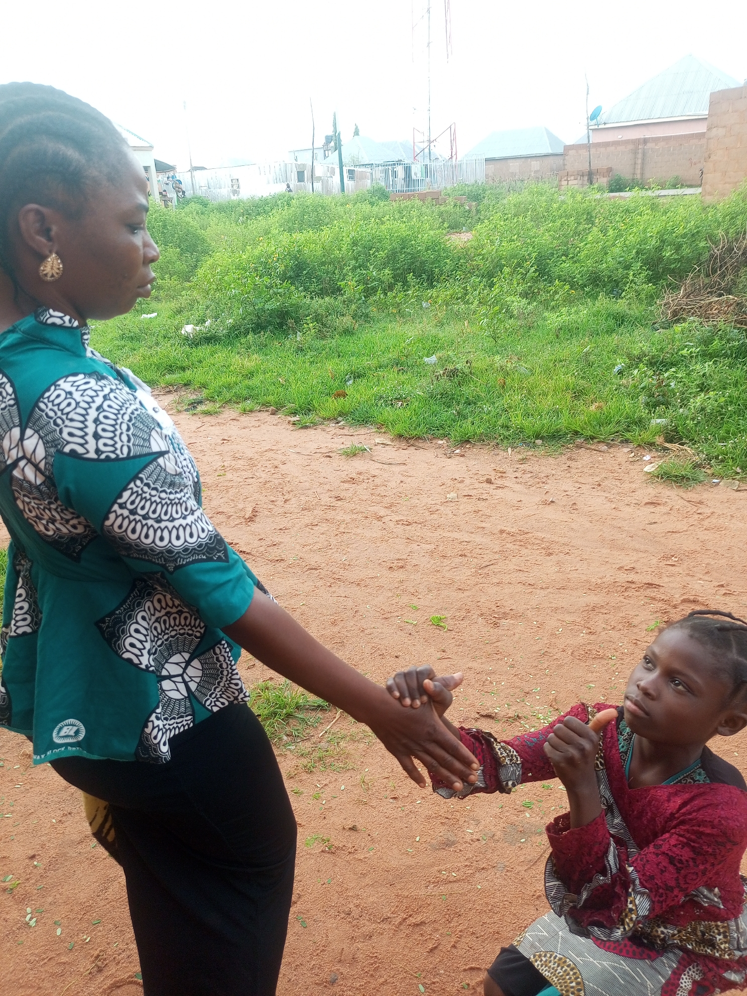 In Tempure, Nigeria, an adult woman, seen in profile and wearing a green and white patterned top and black skirt, extends her right hand. Facing her is a child, kneeling and wearing a red and black patterned garment. The child is reaching up with their right hand, holding what appears to be a piece of food, possibly a carrot, towards the woman's hand. Their left hand is held near the woman's left hand, suggesting a potential handshake or interaction. The background consists of an outdoor environment with sparse, dry vegetation and sandy ground. In the distance, buildings and structures, some with corrugated metal roofs, are visible, along with a radio tower. The sky is overcast.
