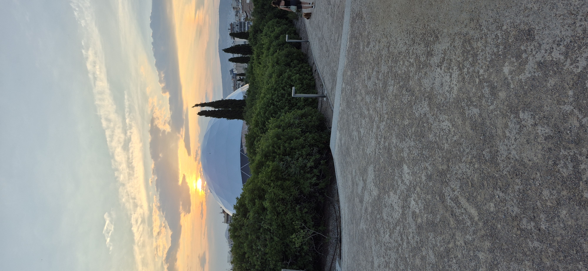 The image shows a geodesic dome structure, likely the Stavros Niarchos Foundation Cultural Center, in Kallithea, Greece, during sunset. The dome is a large, white, semi-spherical building with a visible geometric framework. It is surrounded by lush green trees and bushes. In the foreground, there is a pathway made of gravel and concrete. A person is walking on the pathway with a dog on a leash. The sky is filled with soft orange and yellow hues of the setting sun, with scattered clouds. The overall scene suggests a tranquil evening in an urban park or public space.