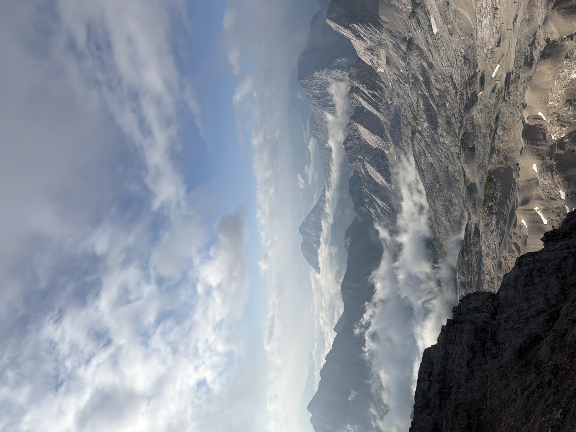 The image depicts a high-altitude mountain landscape, likely in or near Grainau, Germany, given the context. The foreground is dominated by a dark, rocky cliff face. Beyond this, a vast expanse of rugged, grey mountains stretches into the distance, some peaks partially obscured by a layer of white clouds that float between the ranges. The sky above is a mix of blue and white clouds, with sunlight filtering through, creating a bright, airy atmosphere. The terrain appears barren and rocky, with sparse vegetation in some areas and patches of snow on higher slopes. The overall scene conveys a sense of natural grandeur and isolation.