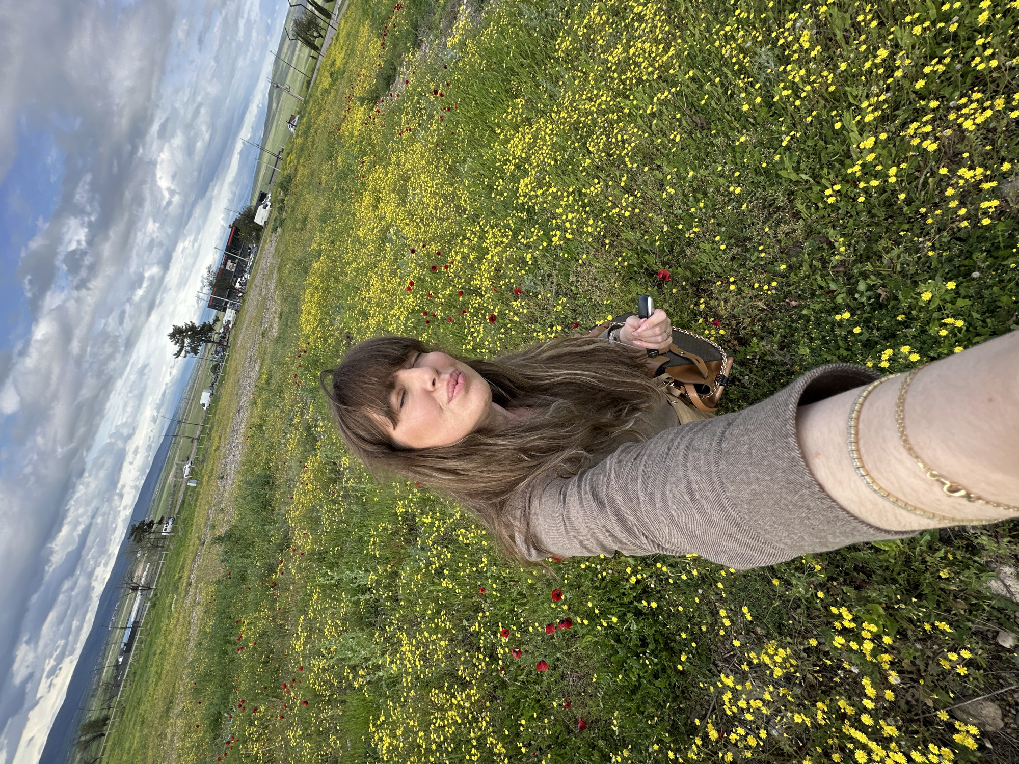 A selfie depicts a woman with long, brown hair, and bangs, pouting her lips. She is positioned in a field filled with yellow wildflowers and sporadic red poppies. The photo is taken from a low angle, with the camera held at arm's length, capturing her face and arm in the foreground. She is wearing a brown jacket and at least one gold bracelet on her wrist, and holding a small object. The background includes a cloudy sky, distant mountains, and what appears to be a roadside area with utility poles, buildings, and parked vehicles. This scene could potentially be located near Marneuli, Georgia, given the landscape and vegetation.