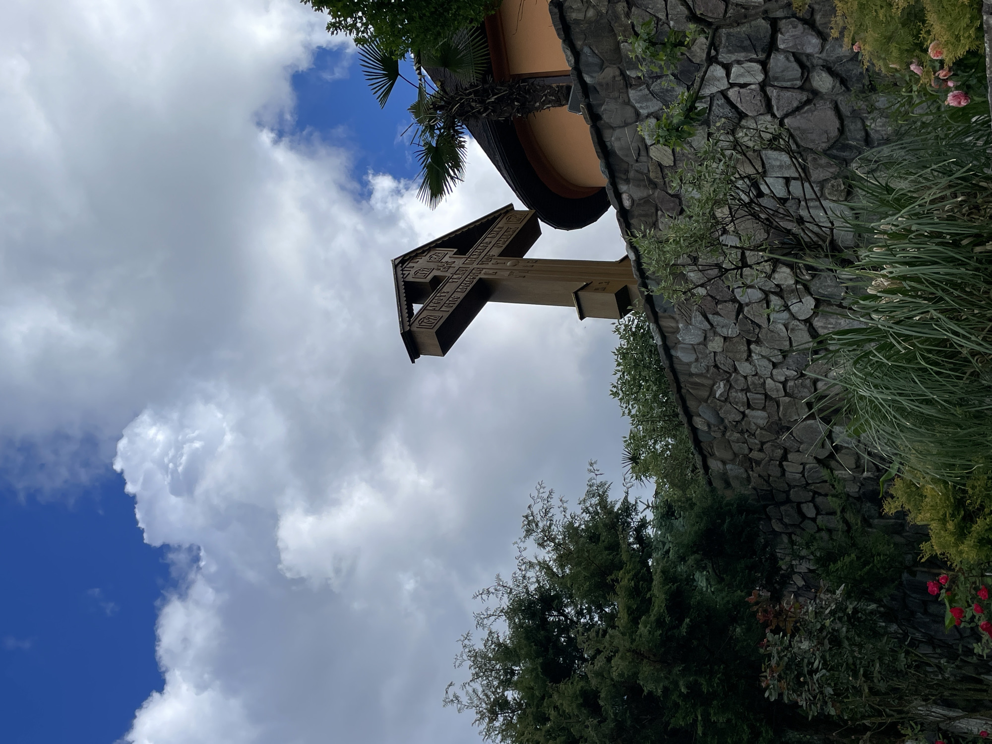 A low-angle shot depicts a carved wooden cross standing against a cloudy sky, located in Solokh-Aul, Russia. The cross, situated near a stone-clad structure and surrounded by lush greenery including trees and shrubs, appears to be part of a religious or commemorative monument. The background shows a mix of blue sky and white clouds.
