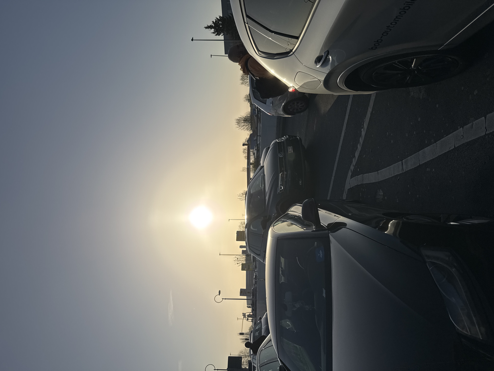 The image shows a parking lot scene in Dortmund, Germany during daylight. Several cars are parked. A person is standing next to a white car, possibly entering or exiting. The sun is high in the sky, creating a bright glare. Streetlights are visible in the background along with buildings. The overall scene suggests an outdoor environment on a sunny day, with a focus on automobiles.