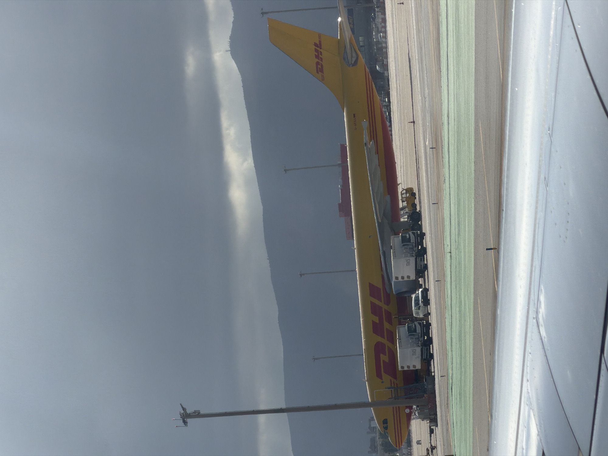 A DHL cargo airplane is parked on the tarmac at an airport. The airplane is predominantly yellow with red accents and the DHL logo is visible on the fuselage and tail. Several white trucks and a small white van are positioned near the airplane, likely for cargo loading or unloading. In the background, rolling hills and mountains can be seen, partially obscured by a cloudy, overcast sky. The foreground shows a section of an aircraft wing, suggesting the photo was taken from inside another airplane. The scene appears to be at Barcelona-El Prat Airport, a major international airport serving the city of Barcelona, Spain. The lighting suggests it is daytime, but the clouds create a somewhat subdued atmosphere. No people are clearly visible in the frame.