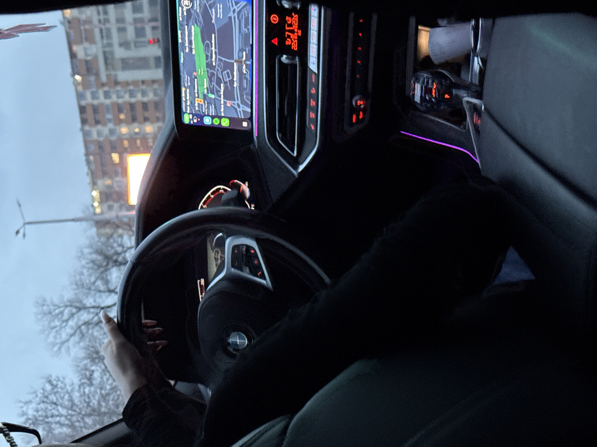 The image shows the interior of a BMW car. The driver is holding the steering wheel with one hand. The car's dashboard is visible, including the navigation screen displaying a map, the climate control panel, and the ambient lighting. Outside, there is a view of a tall building. A street light and bare trees are also visible through the windshield. The location could be Plovdiv, Bulgaria, given the prompt.